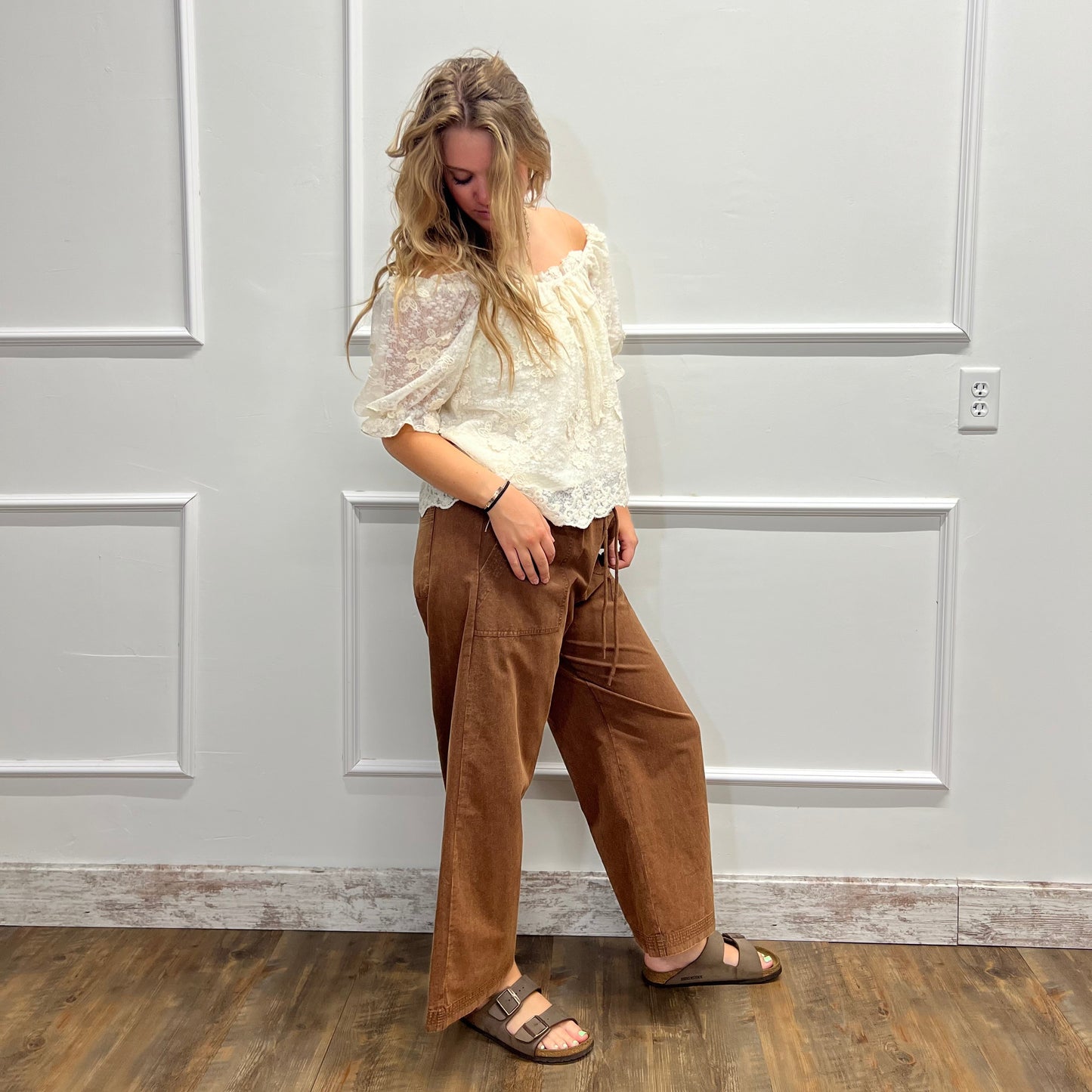 A woman stands in a room with white paneled walls and a wood floor, wearing a cream lace off-the-shoulder blouse, brown wide-leg corduroy pants, and brown slide sandals.