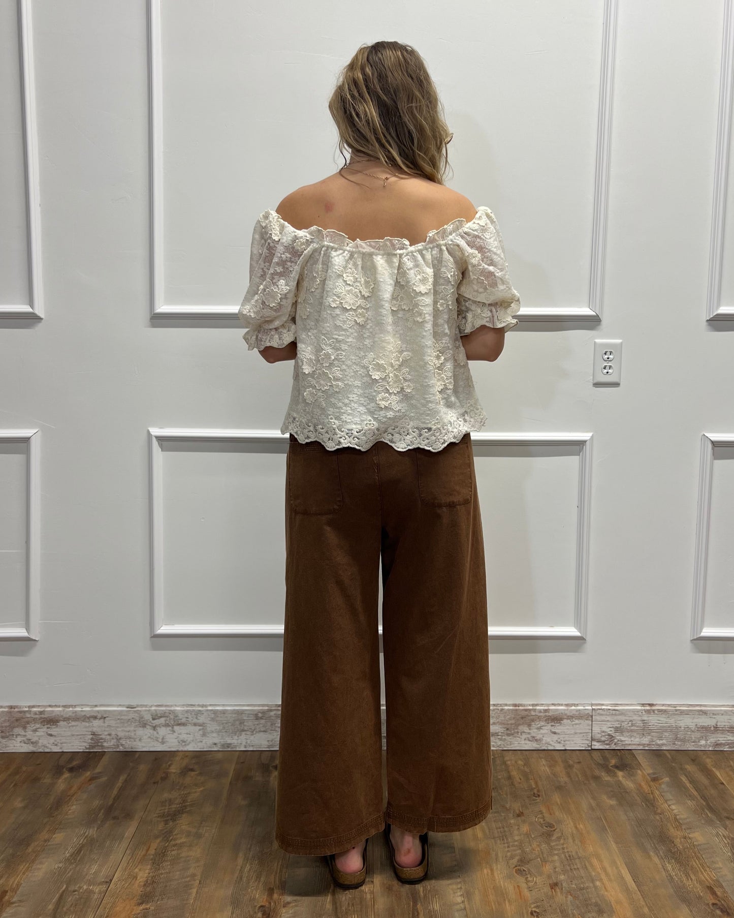 A woman viewed from behind wearing an off-the-shoulder white lace top and brown wide-leg pants, standing against a white paneled wall on a wood floor.