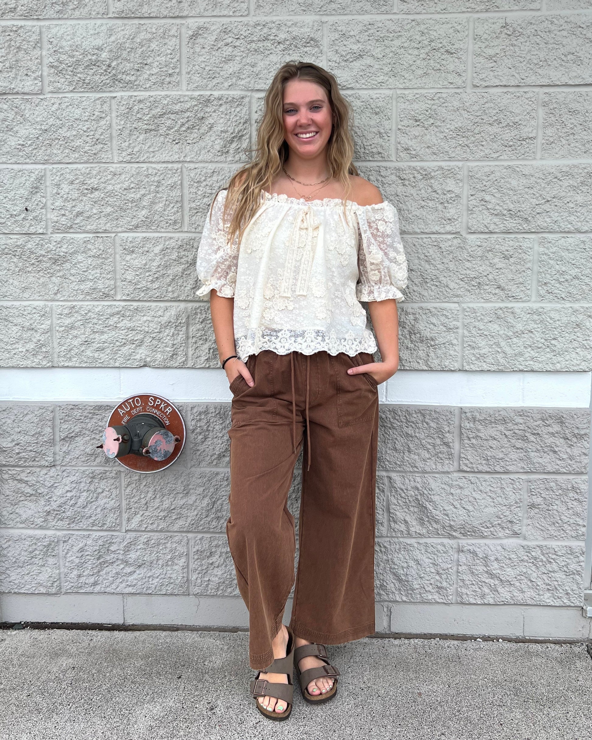 A woman stands outdoors against a light gray stone wall wearing a cream lace-trim off-the-shoulder top, brown wide-leg pants, and black sandals.