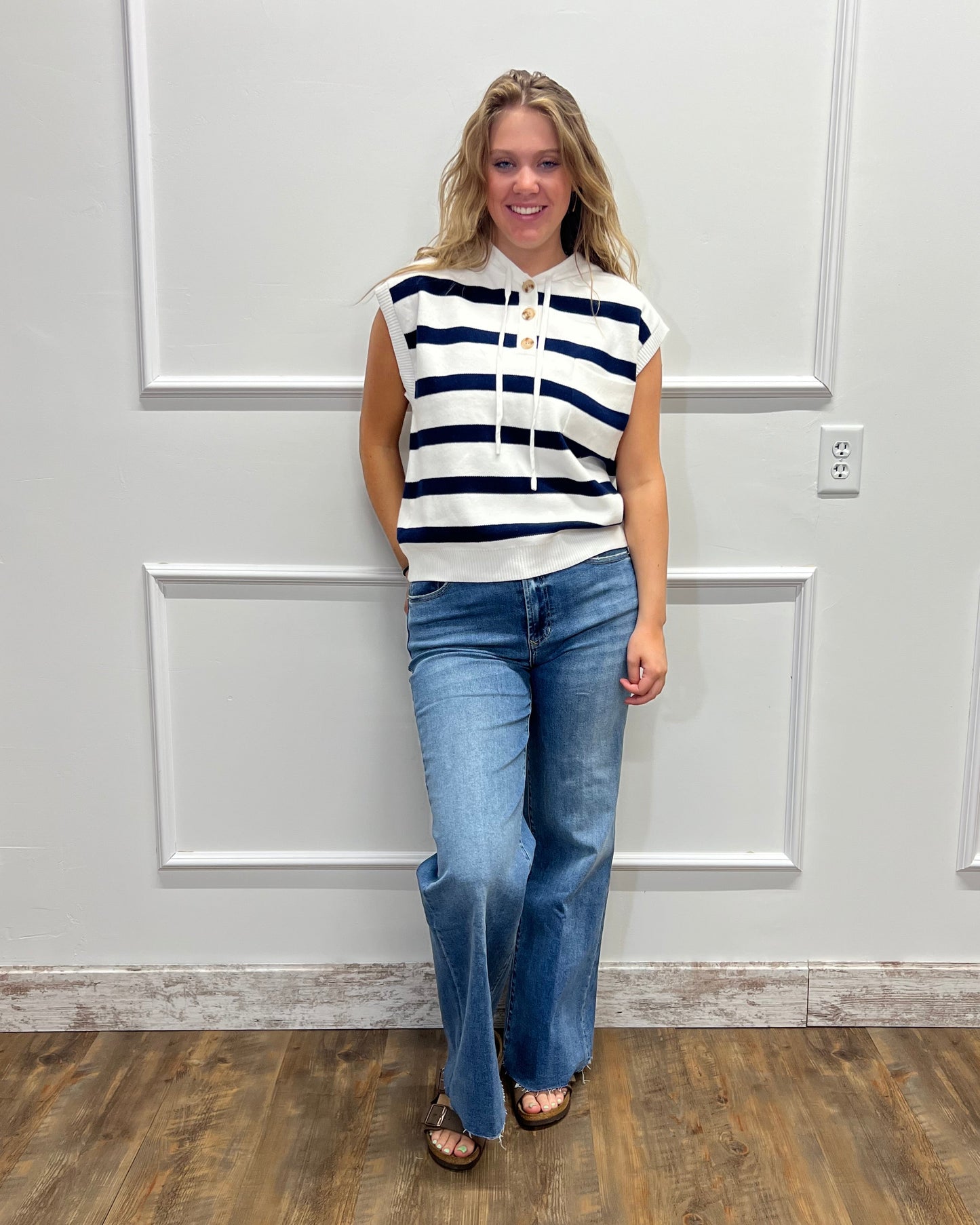 A woman wearing a navy and white striped sleeveless top, blue jeans, and brown sandals standing in front of a white paneled door with a wood-look floor visible on the right.