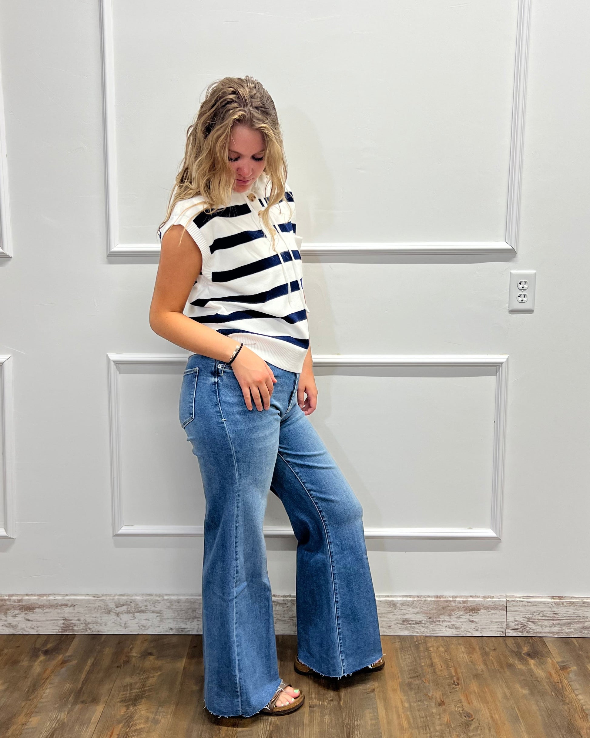 A woman stands against a white paneled wall wearing a black and white horizontally striped sleeveless top and blue flare jeans.