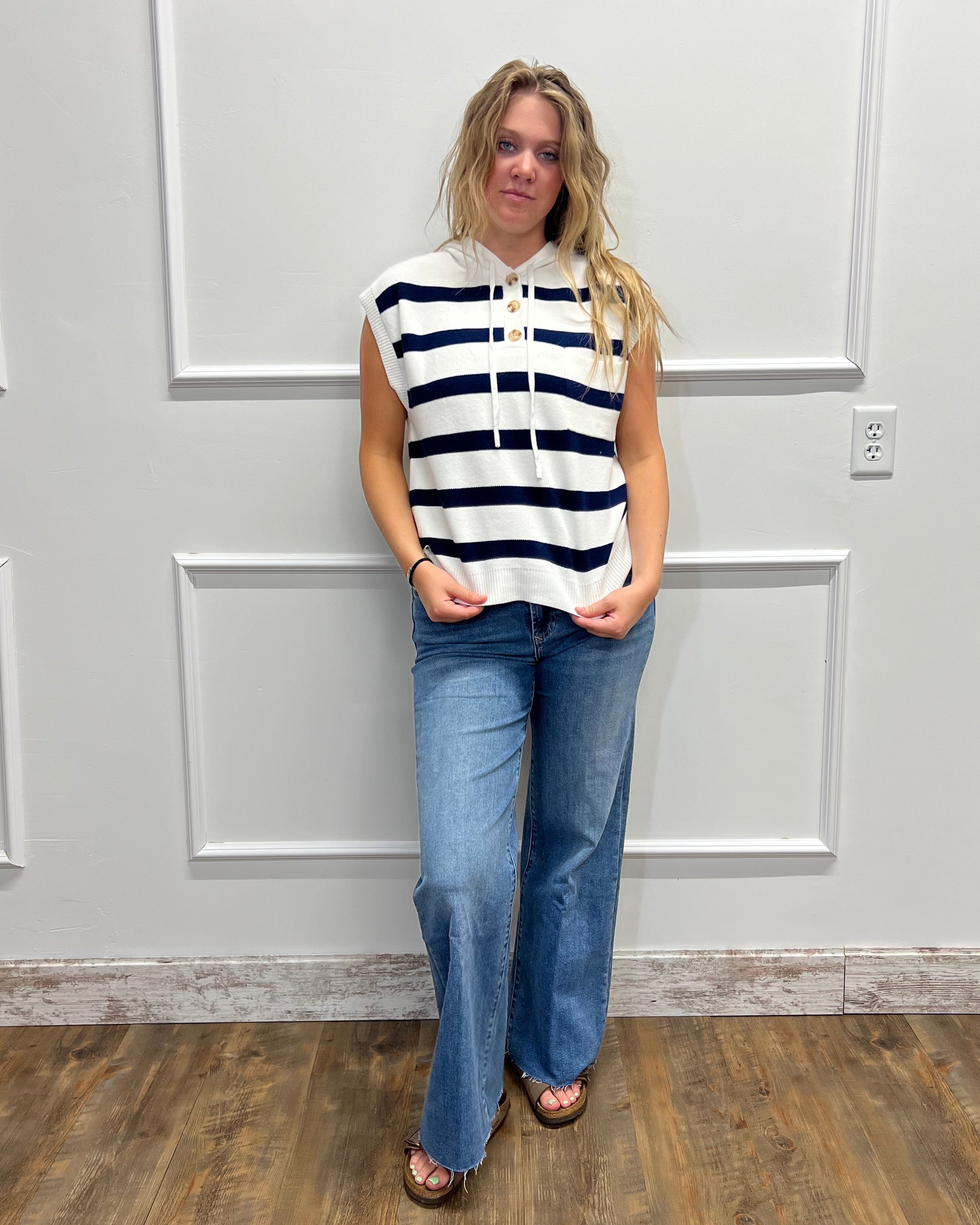 A woman modeling a sleeveless black and white striped top with a drawstring, paired with blue jeans and brown sandals, standing in front of a white paneled door.