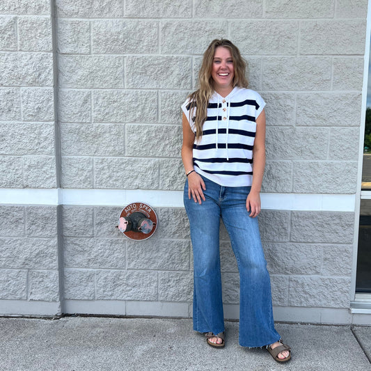 A woman wearing a black and white striped short-sleeve top, blue jeans, and brown sandals stands outdoors against a light gray textured wall.