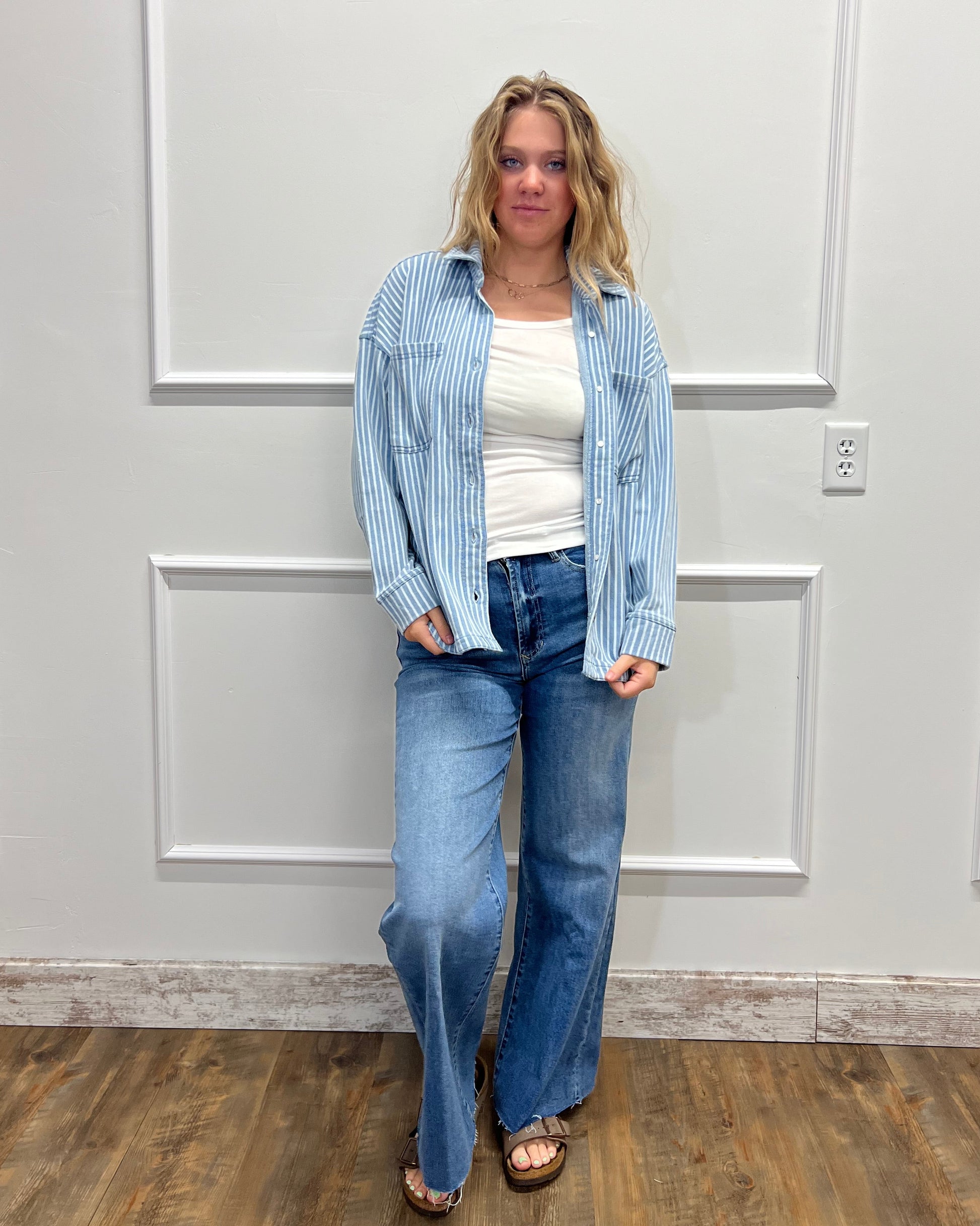 A woman modeling a light blue and white striped button-up shirt over a white top, paired with blue jeans and brown sandals, standing in front of a white paneled door.