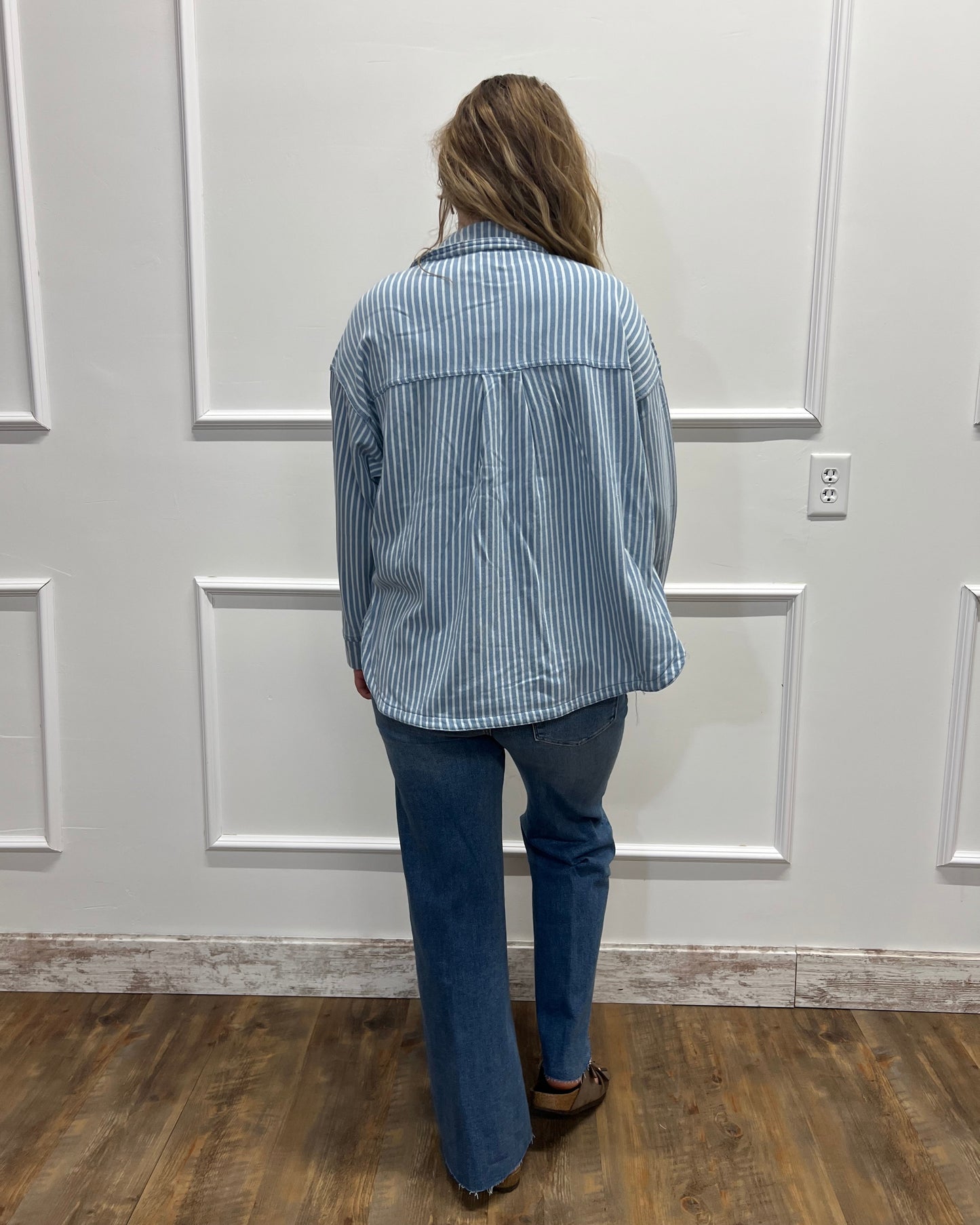 A person viewed from behind wearing a light blue and white vertically striped button-up shirt and dark blue jeans, standing in front of a white paneled door on a wood floor.