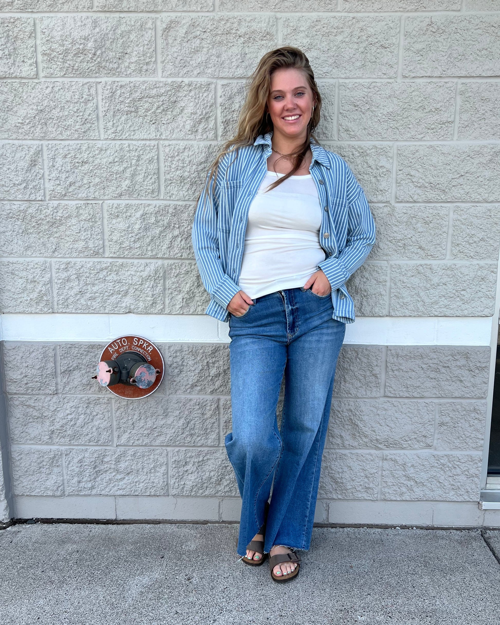 A woman wearing a light blue and white striped button-up shirt, a white t-shirt, and blue jeans, standing on a gray paved sidewalk next to a light gray stone wall.