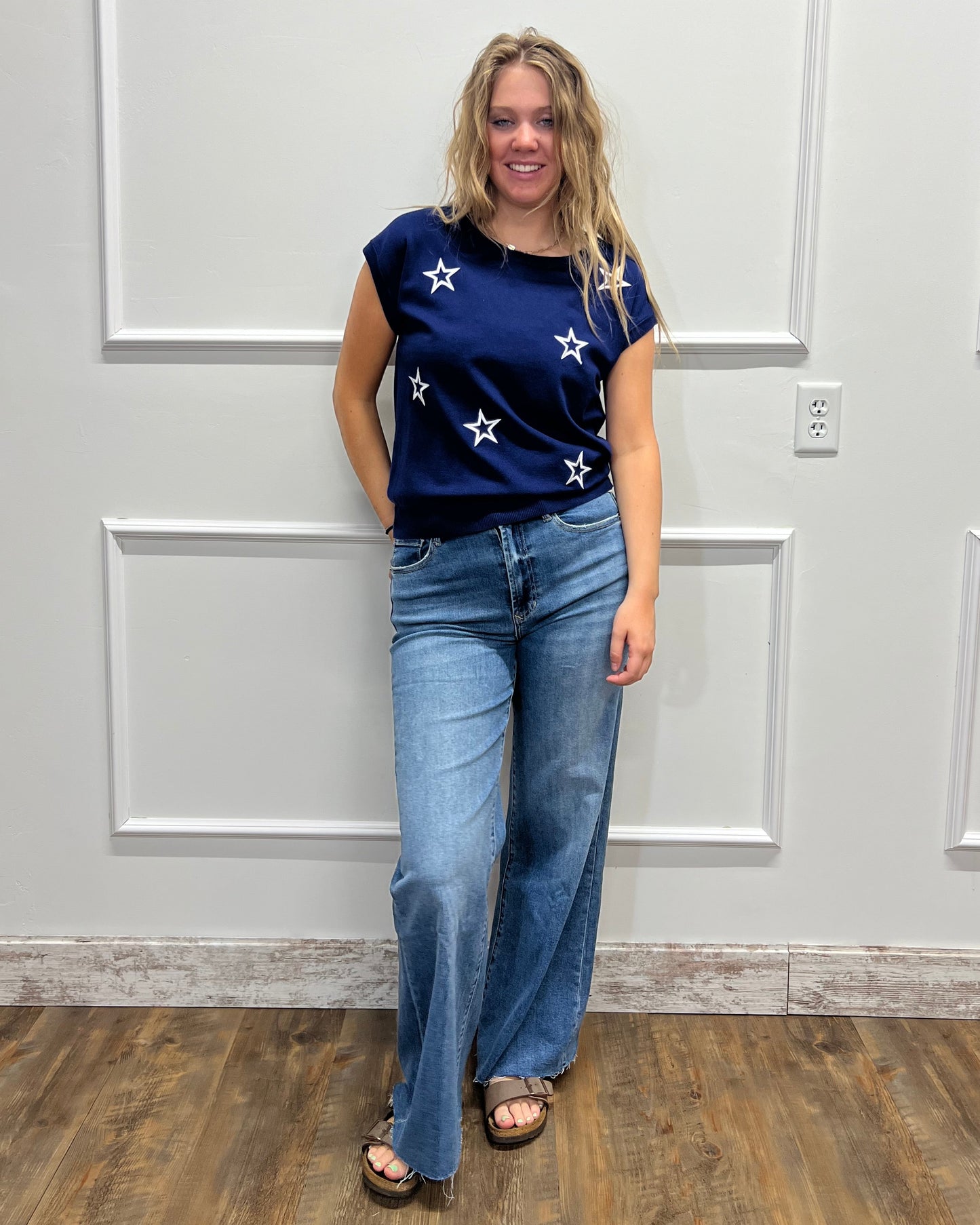 A woman wearing a navy blue short-sleeve top with white star patterns and light blue jeans, standing in front of a white paneled wall.