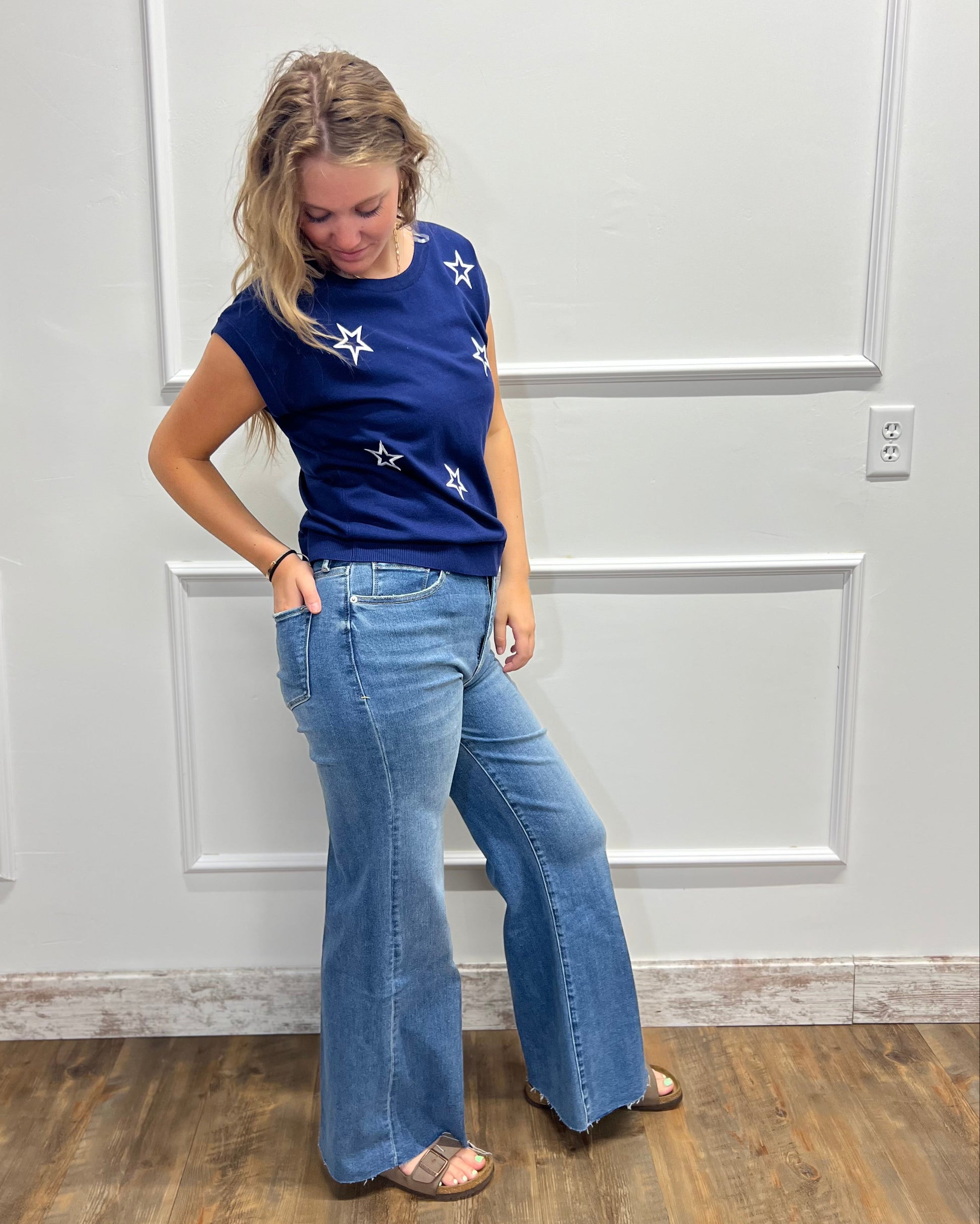 A woman modeling a navy blue short-sleeve top with white star patterns and light-wash blue flare jeans, standing against a white paneled wall.