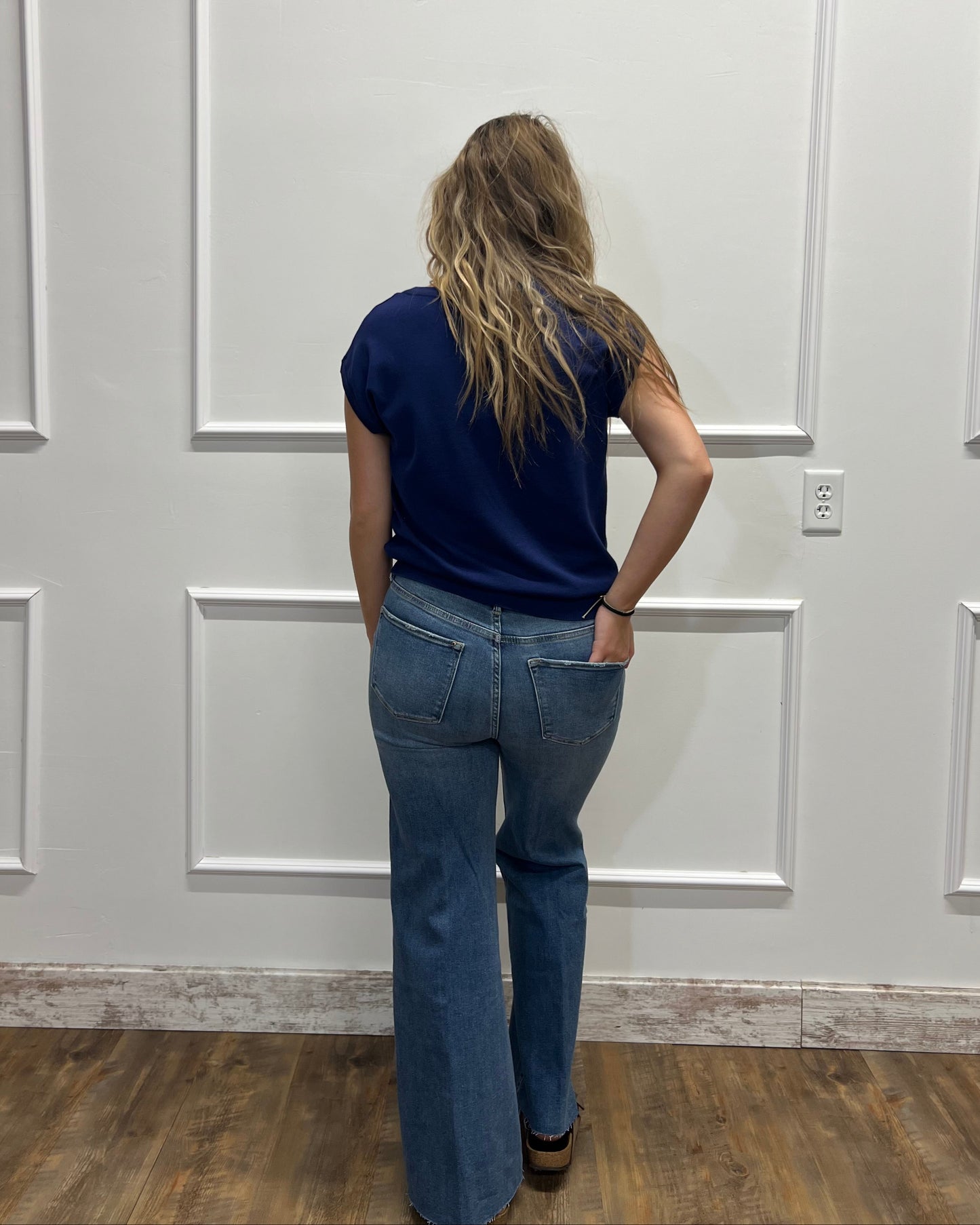 A woman viewed from behind wearing a navy blue short-sleeve t-shirt and blue flare jeans, standing in front of a white paneled wall on a wood floor.