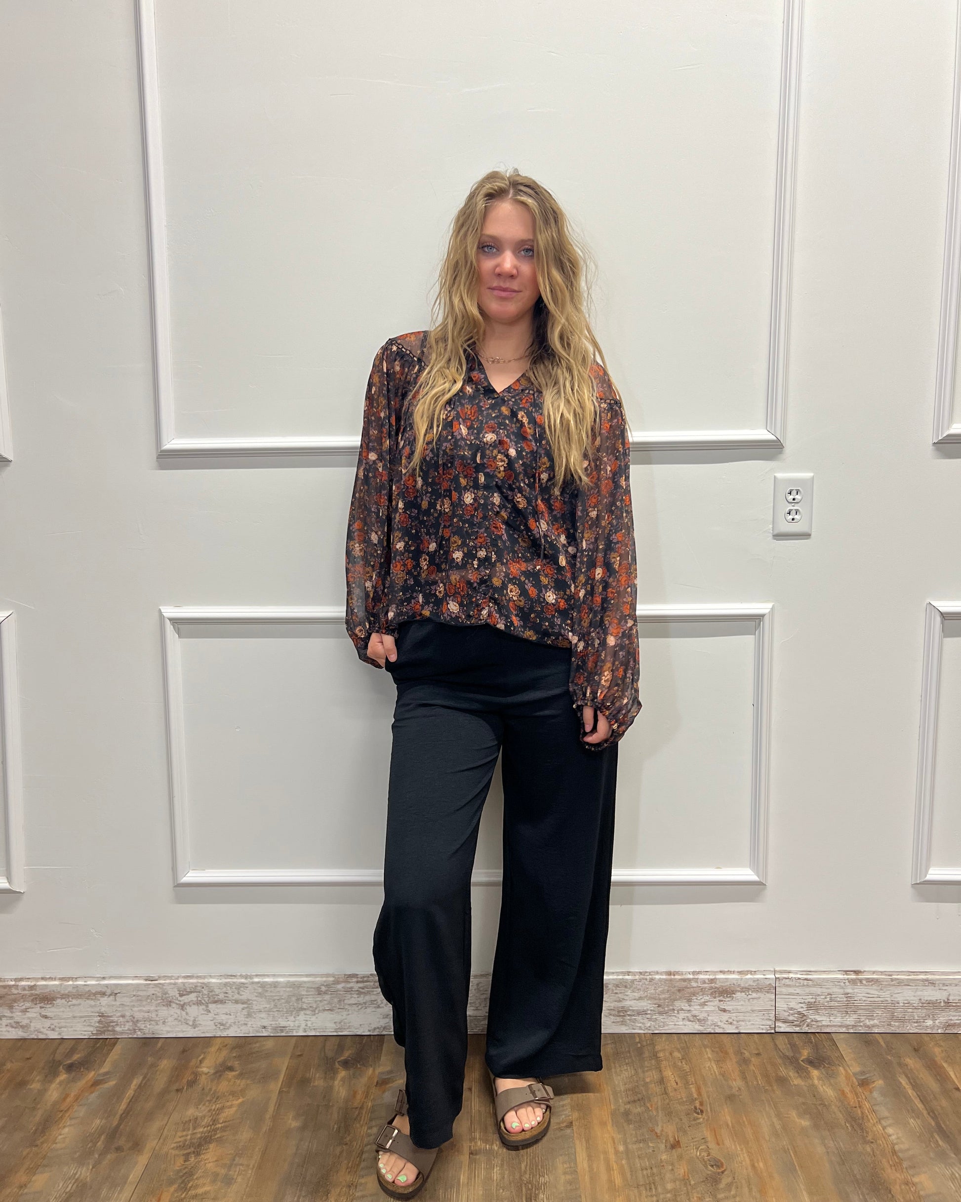 A woman stands in front of a white paneled door wearing a long-sleeve floral patterned blouse, black wide-leg pants, and brown sandals.