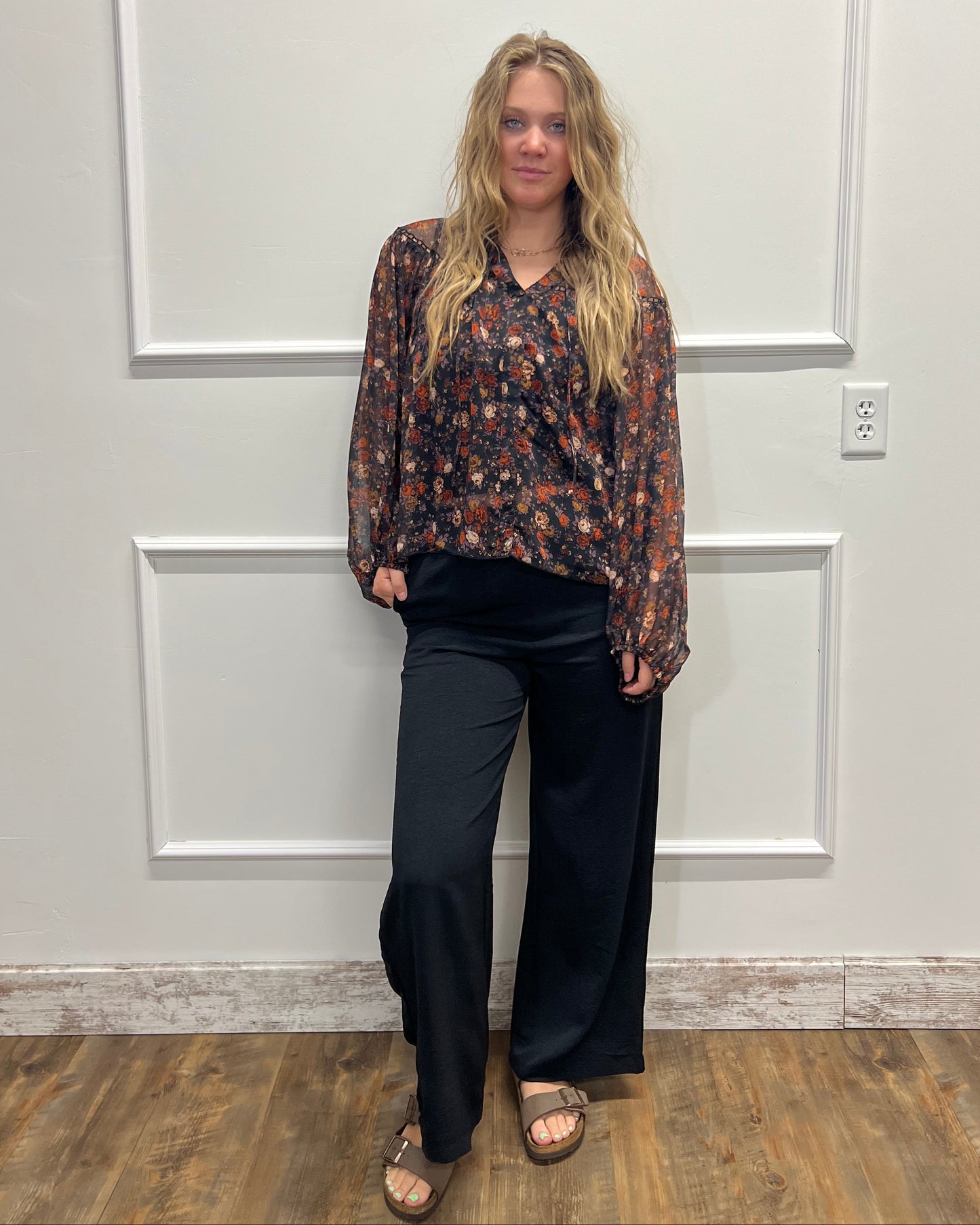 A woman stands in front of a white paneled door wearing a long-sleeve floral patterned blouse, black wide-leg pants, and brown sandals.