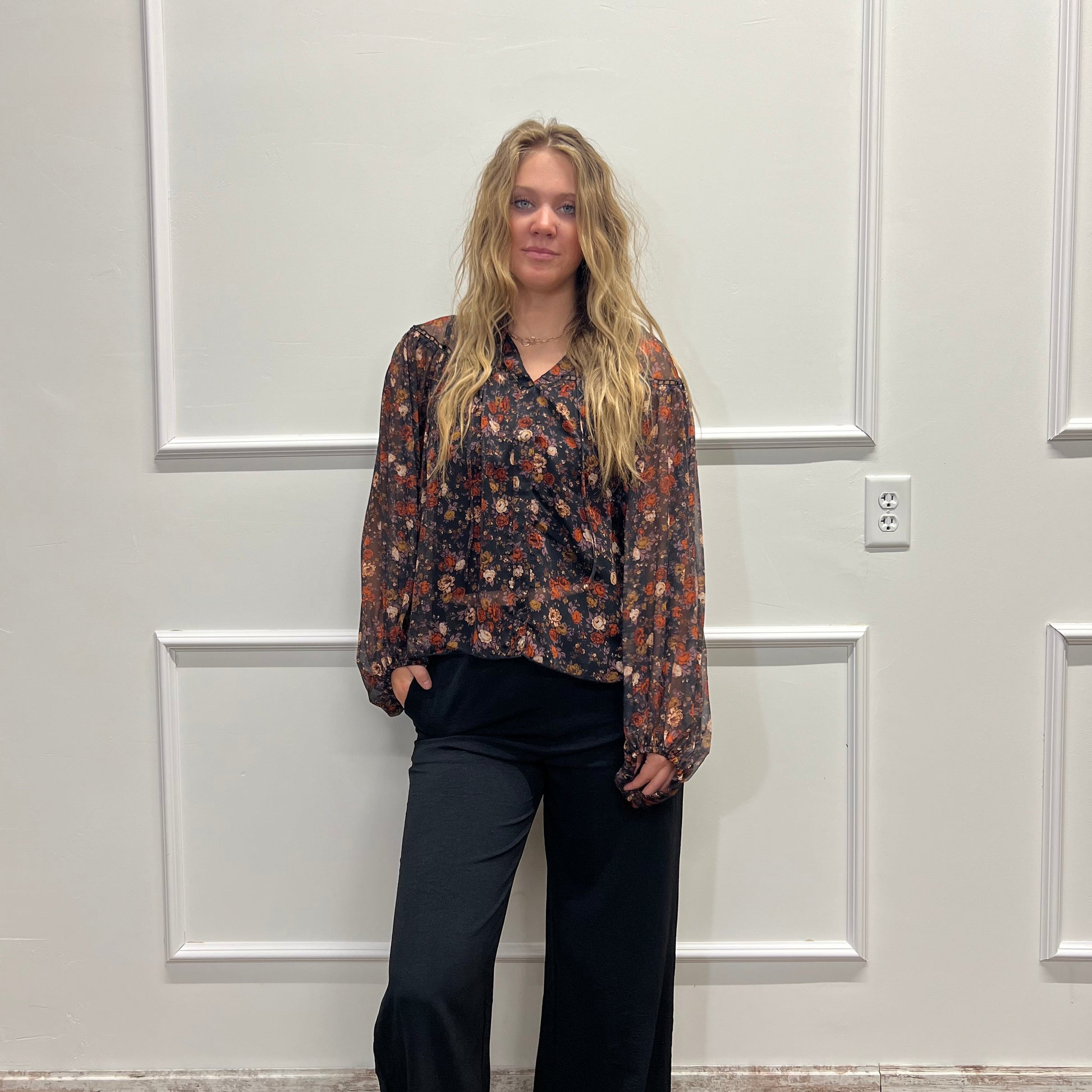 A woman stands in front of a white paneled door wearing a dark floral patterned long-sleeve top and black wide-leg pants.