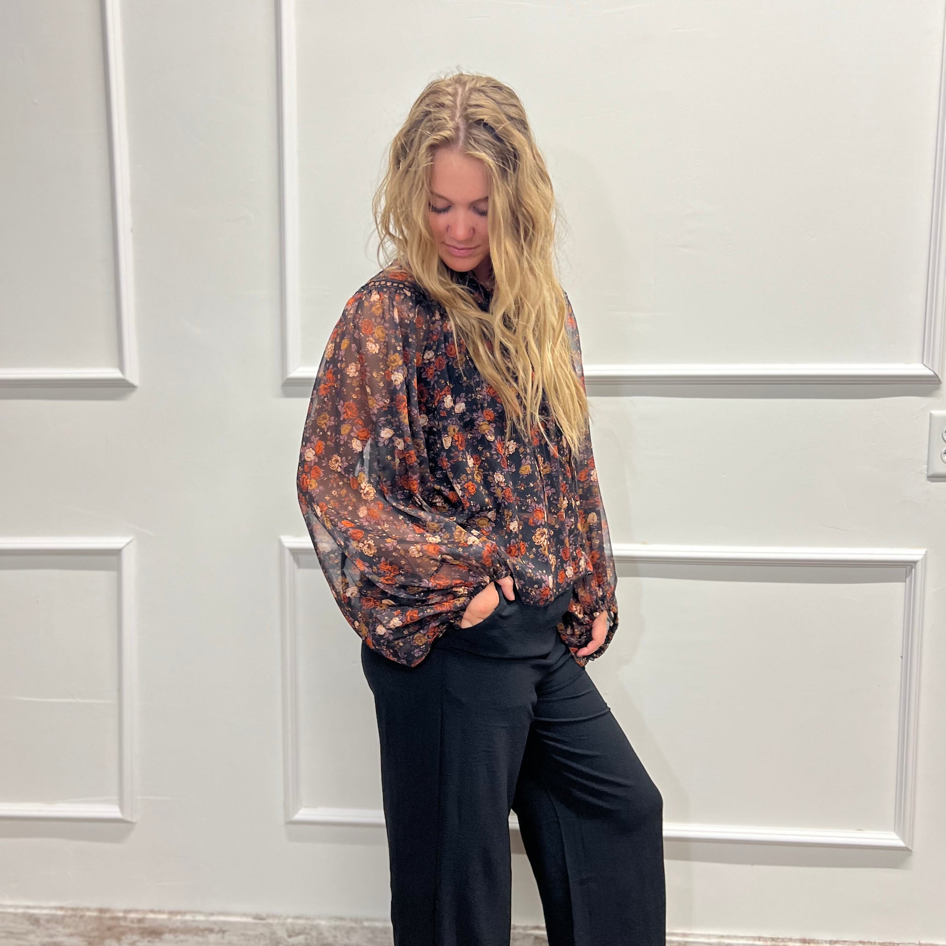 A woman with blonde hair stands against a white paneled wall wearing a loose, long-sleeve floral blouse in dark tones with black wide-leg pants.