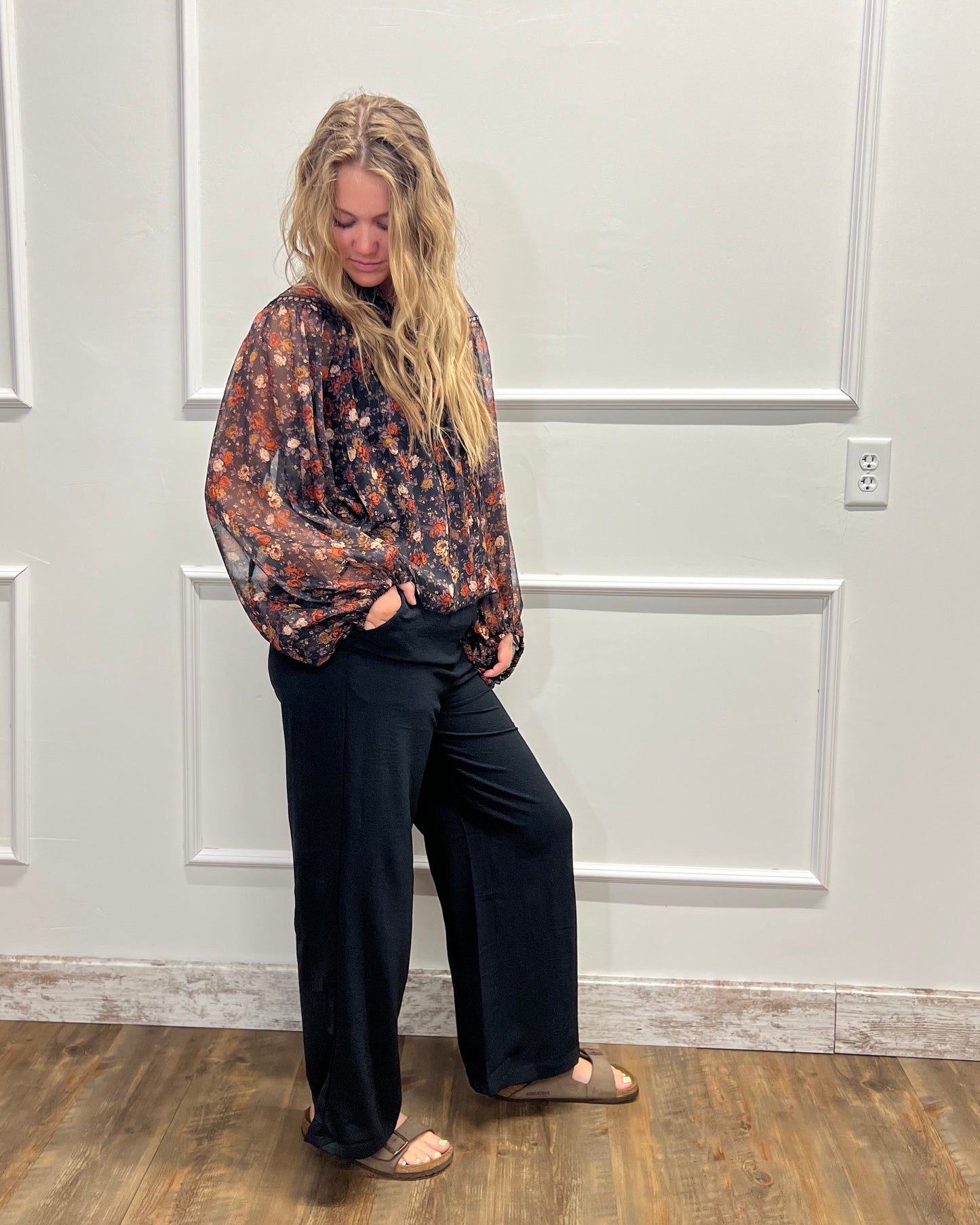 A woman stands in a room with white paneled walls and a wood floor, wearing a loose, long-sleeve floral blouse in dark tones with wide-leg black pants and tan flats.