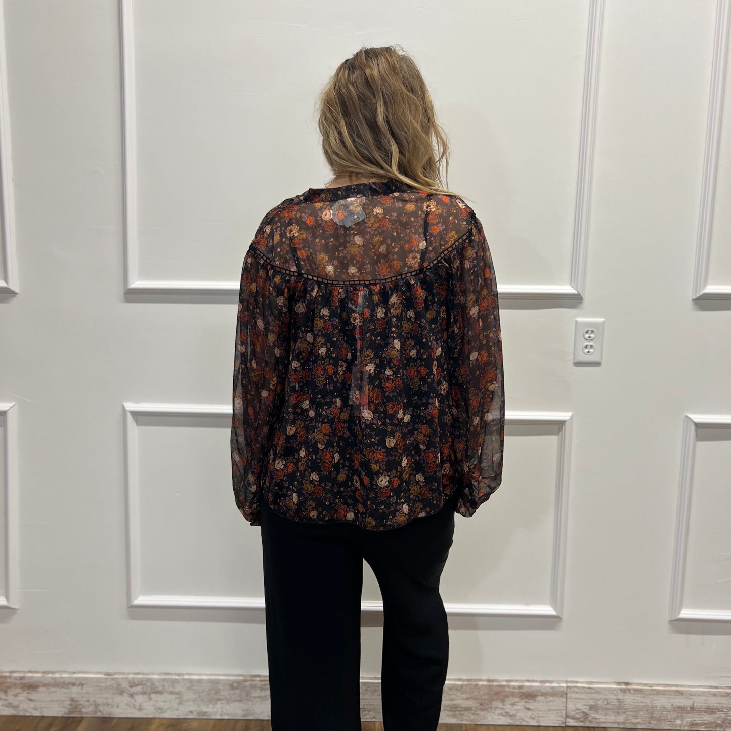 A woman viewed from behind wearing a sheer long-sleeve button-up blouse with a dark brown and black floral pattern, paired with black pants, standing in front of a white paneled wall.