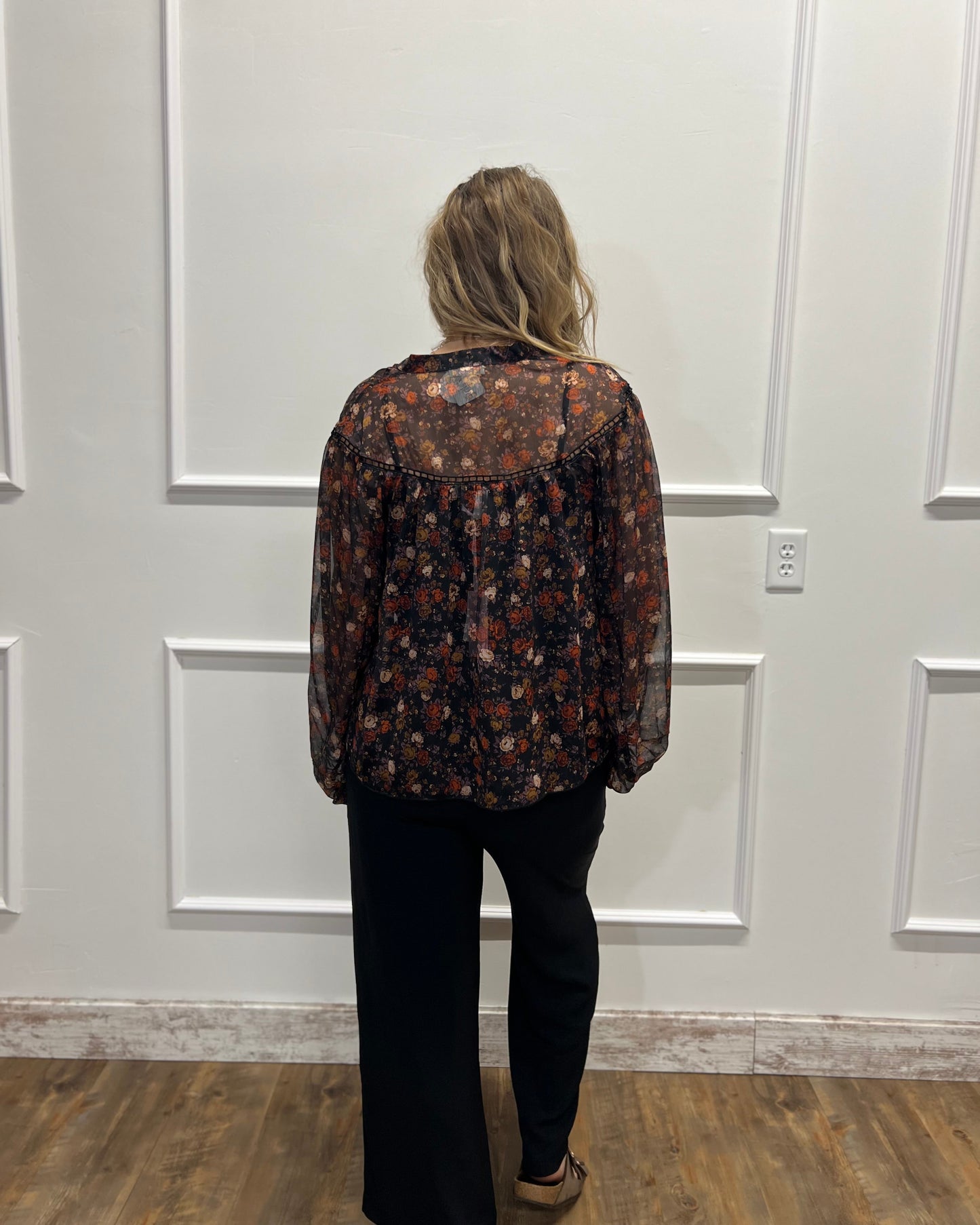 A woman viewed from behind wearing a sheer long-sleeve button-up blouse with a dark brown and rust floral pattern, paired with black pants, standing in front of a white paneled wall.