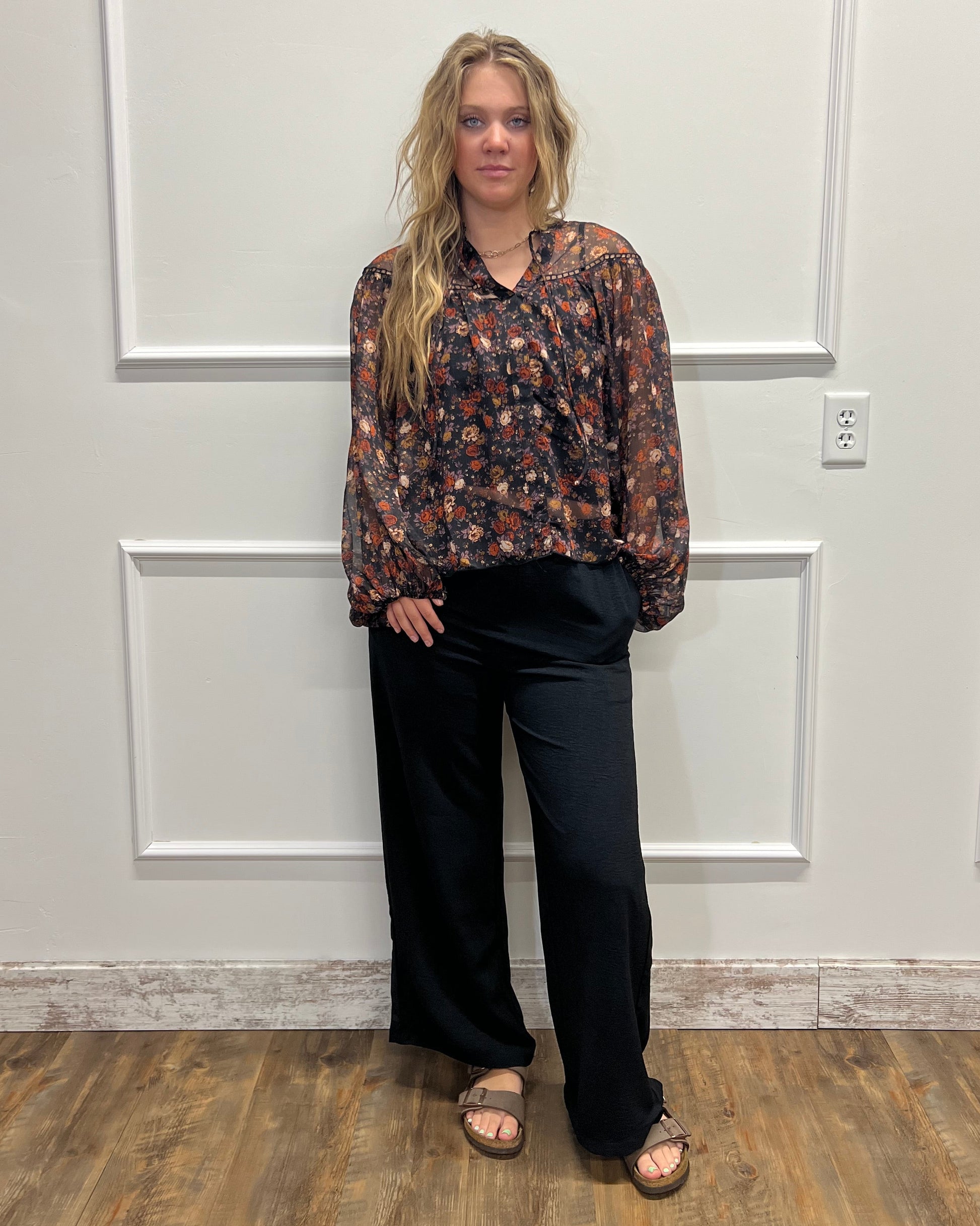 A woman stands in a home interior wearing a dark floral long-sleeve blouse and black wide-leg pants, paired with brown sandals. The photo is oriented sideways, and she is positioned in front of a white paneled door with a wood floor visible on the right.
