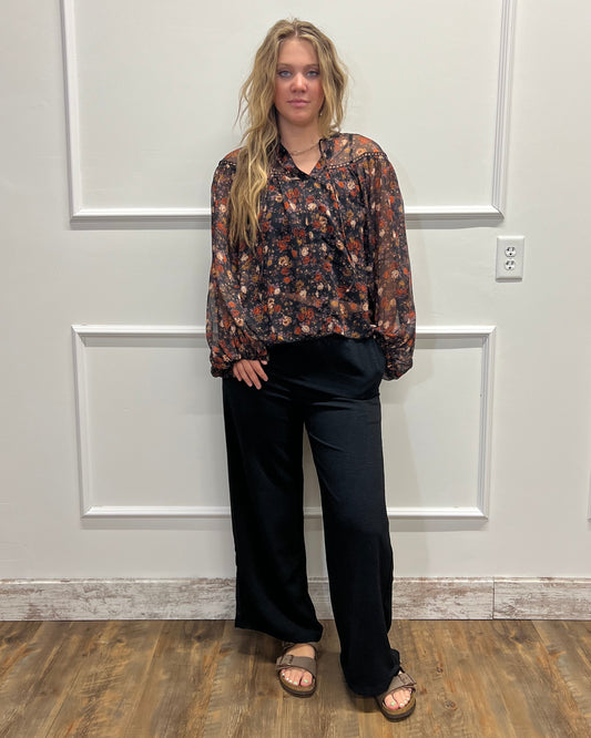 A woman stands in a home interior wearing a dark floral long-sleeve blouse and black wide-leg pants, paired with brown sandals. The photo is oriented sideways, and she is positioned in front of a white paneled door with a wood floor visible on the right.