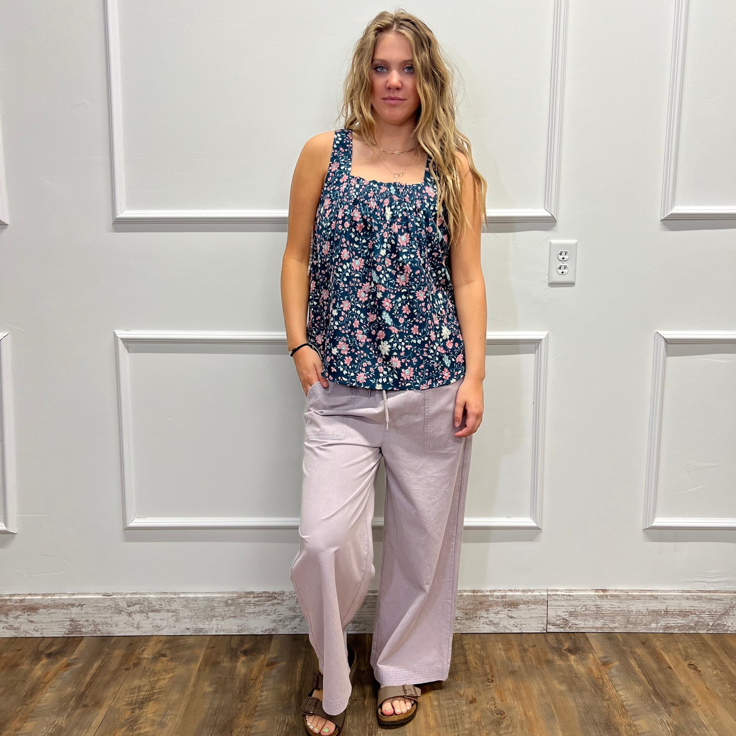 A woman modeling a sleeveless navy blue floral print top and light pink wide-leg pants, standing in front of a white paneled wall with a wood floor visible on the right.