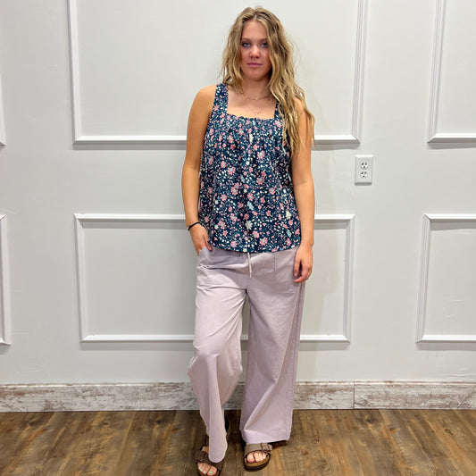 A woman modeling a sleeveless navy blue floral print top and light pink wide-leg pants, standing in front of a white paneled wall with a wood floor visible on the right.