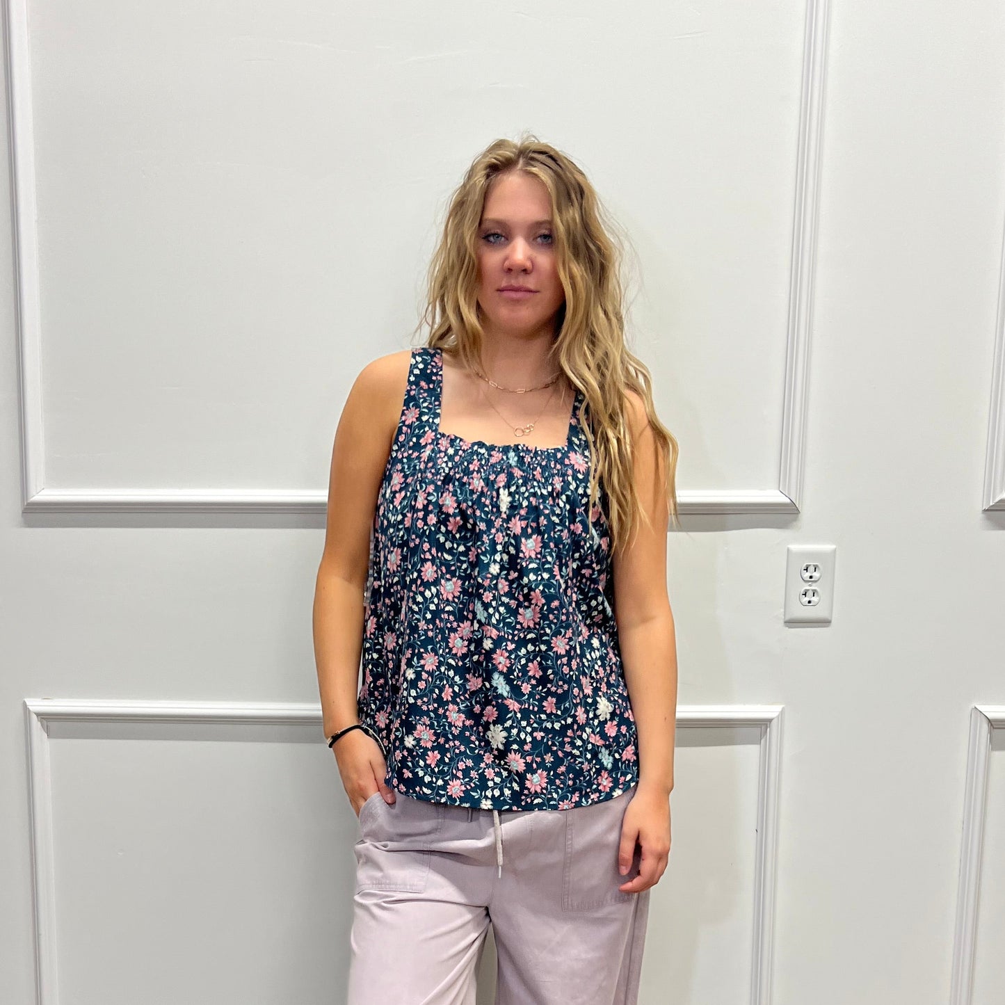 A woman stands in a room wearing a sleeveless floral top and light lavender pants, photographed against a white paneled wall with a wood floor visible on the right.
