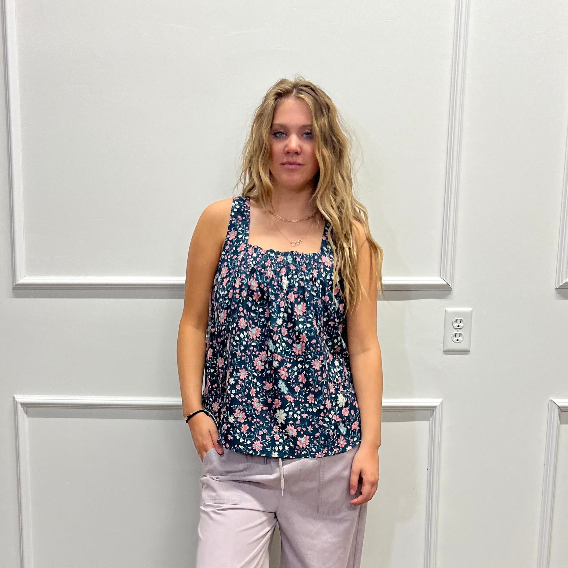 A woman stands in a room wearing a sleeveless floral top and light lavender pants, photographed against a white paneled wall with a wood floor visible on the right.