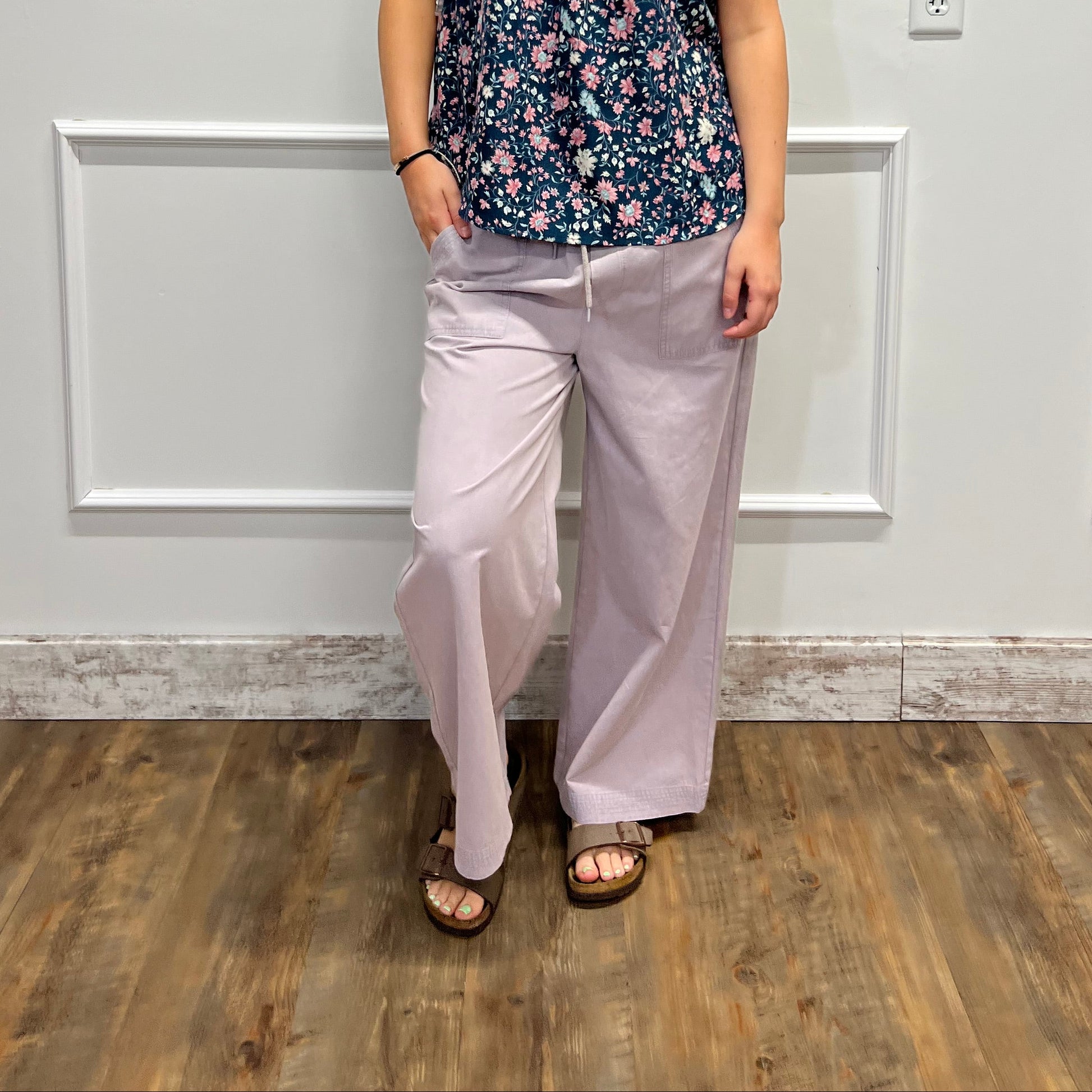 A woman stands in a room wearing a sleeveless floral top and light lavender pants, photographed against a white paneled wall with a wood floor visible on the right.