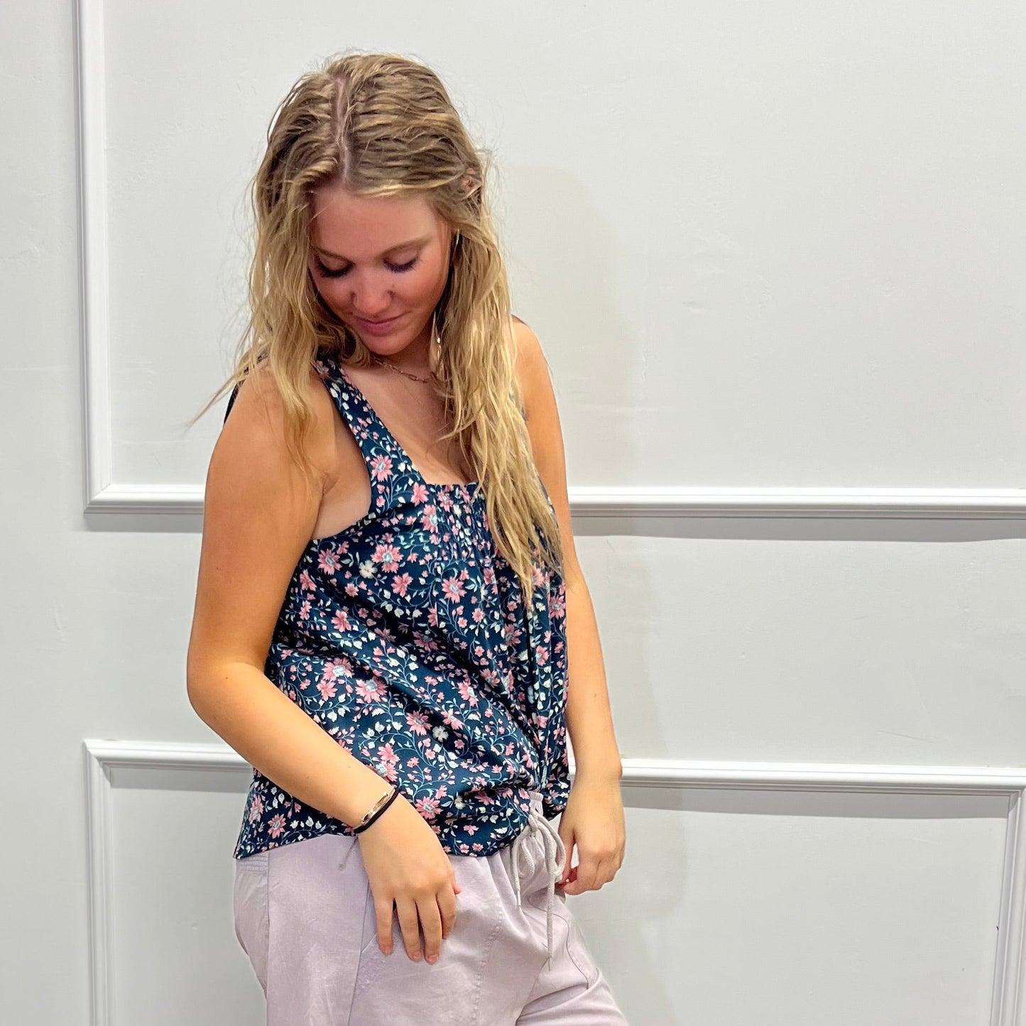 A woman modeling a sleeveless floral tank top and light pink wide-leg pants, standing on a wood floor against a white paneled wall.