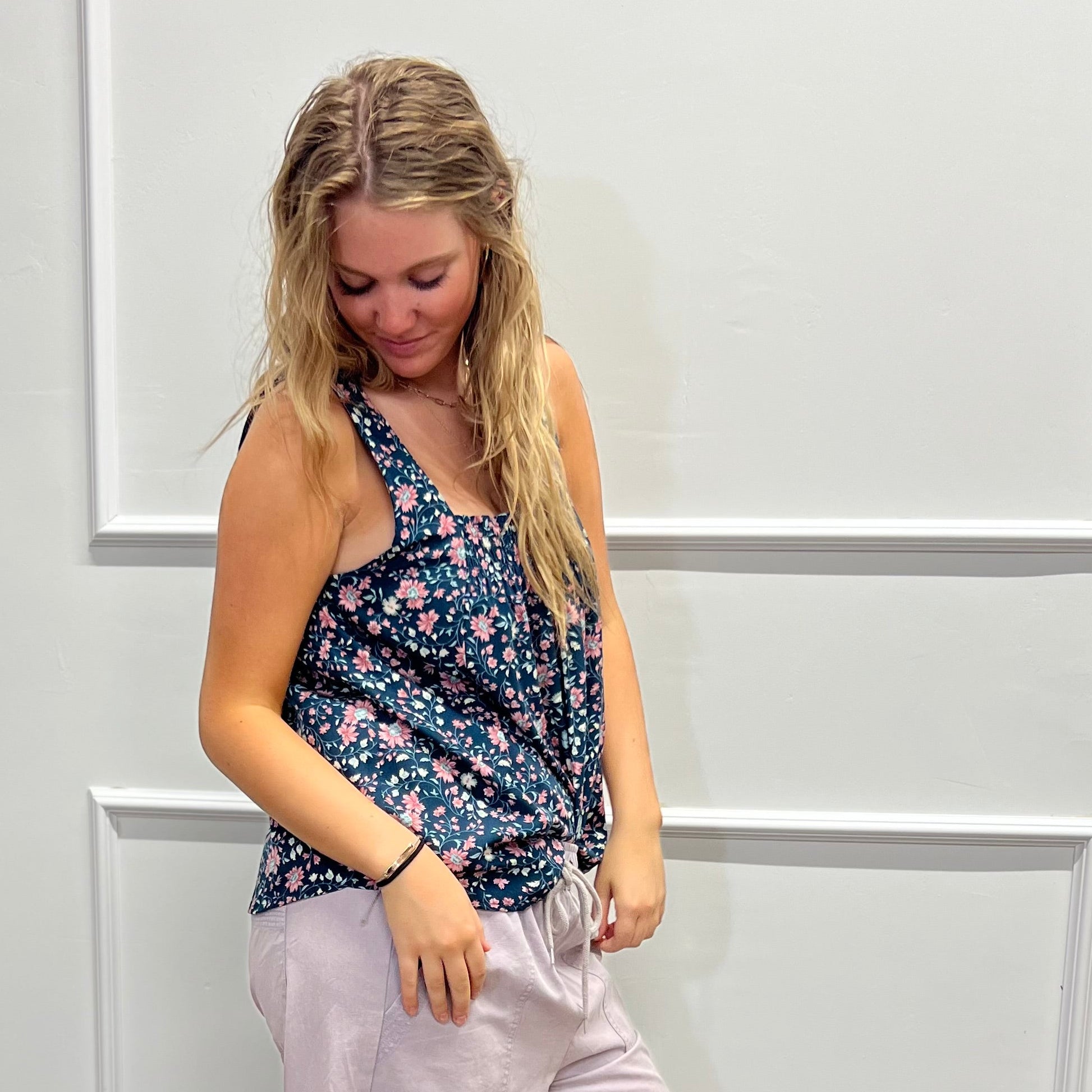 A woman modeling a sleeveless floral tank top and light pink wide-leg pants, standing on a wood floor against a white paneled wall.