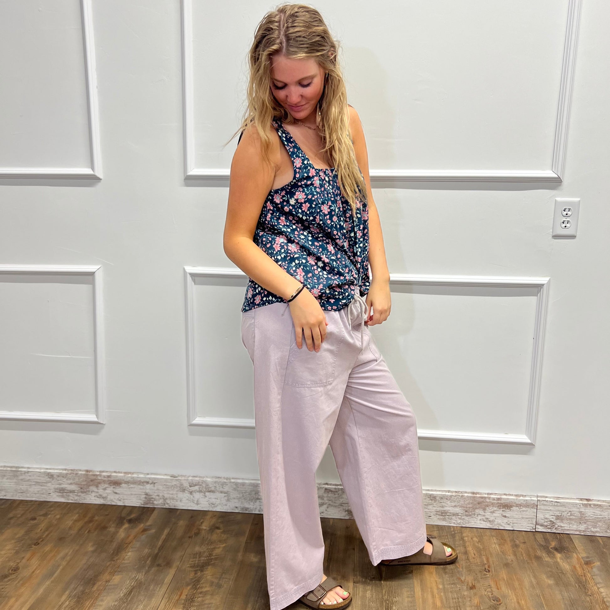 A woman modeling a sleeveless floral tank top and light pink wide-leg pants, standing on a wood floor against a white paneled wall.
