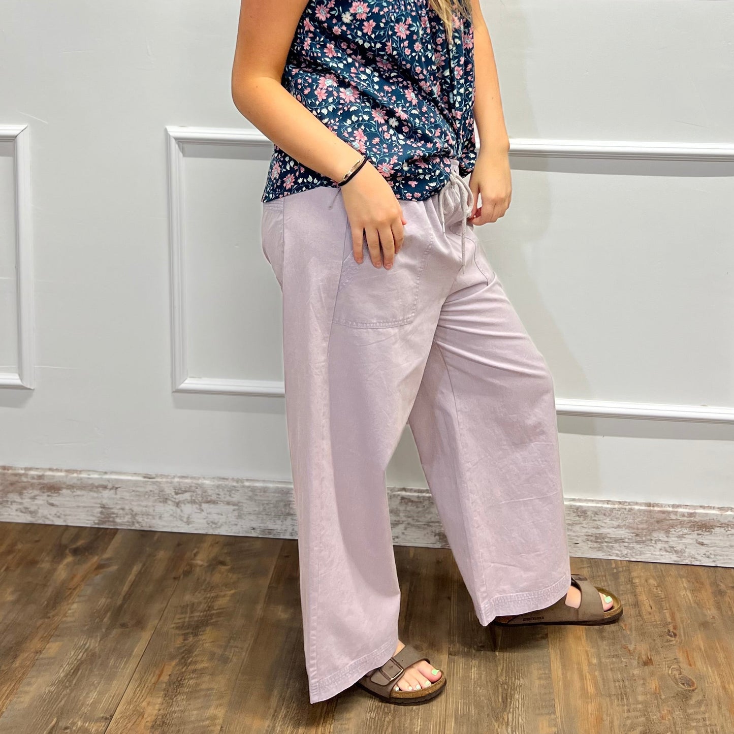 A woman modeling a sleeveless floral tank top and light pink wide-leg pants, standing on a wood floor against a white paneled wall.