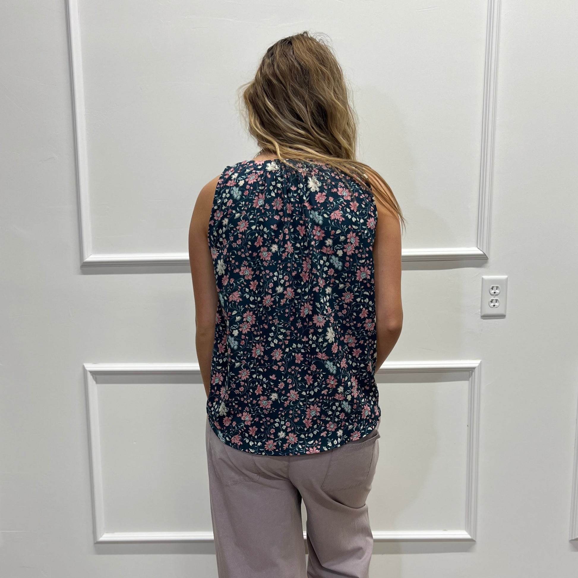 A woman viewed from behind wearing a sleeveless dark floral top and light gray wide-leg pants, standing in front of a white paneled wall on a wood floor.