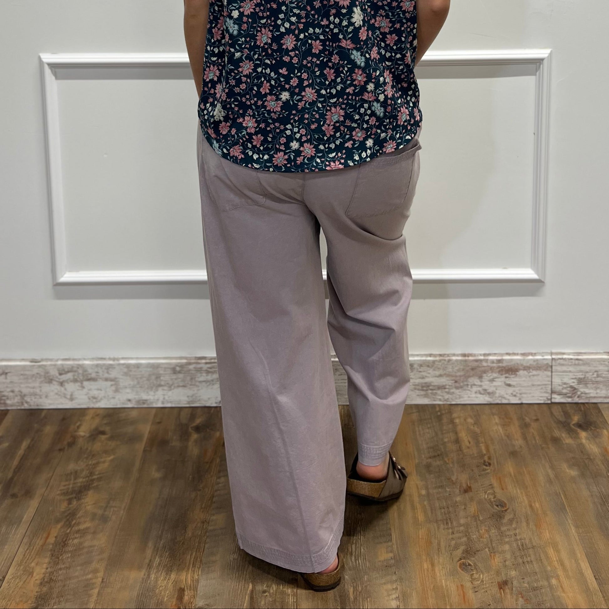 A woman viewed from behind wearing a sleeveless dark floral top and light gray wide-leg pants, standing in front of a white paneled wall on a wood floor.