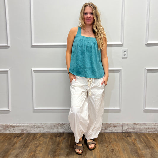 A woman wearing a teal sleeveless top and white pants stands in front of a white paneled wall, with a wood floor visible on the right side.