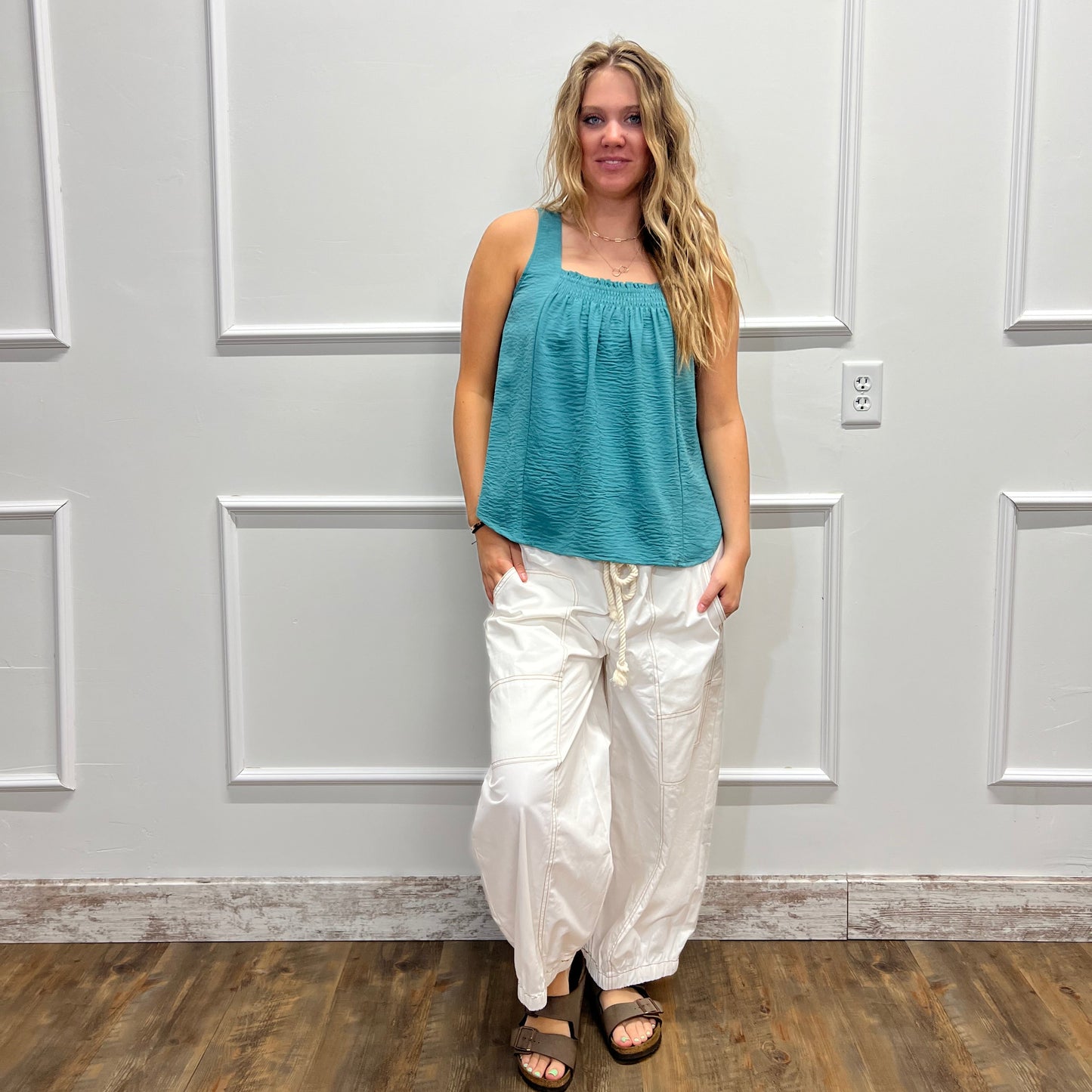 A woman wearing a teal sleeveless top and white pants stands in front of a white paneled wall, with a wood floor visible on the right side.