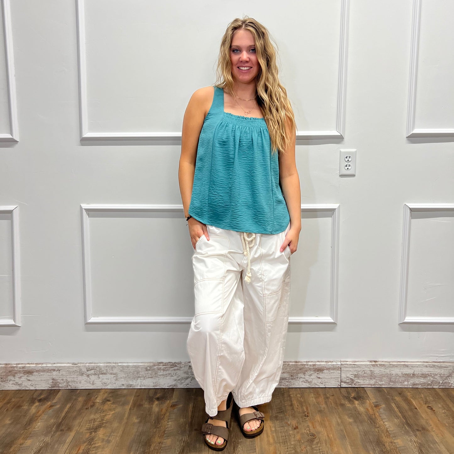 A woman stands indoors against a white paneled wall, wearing a sleeveless teal top and loose white pants with dark sandals. The photo is rotated sideways.
