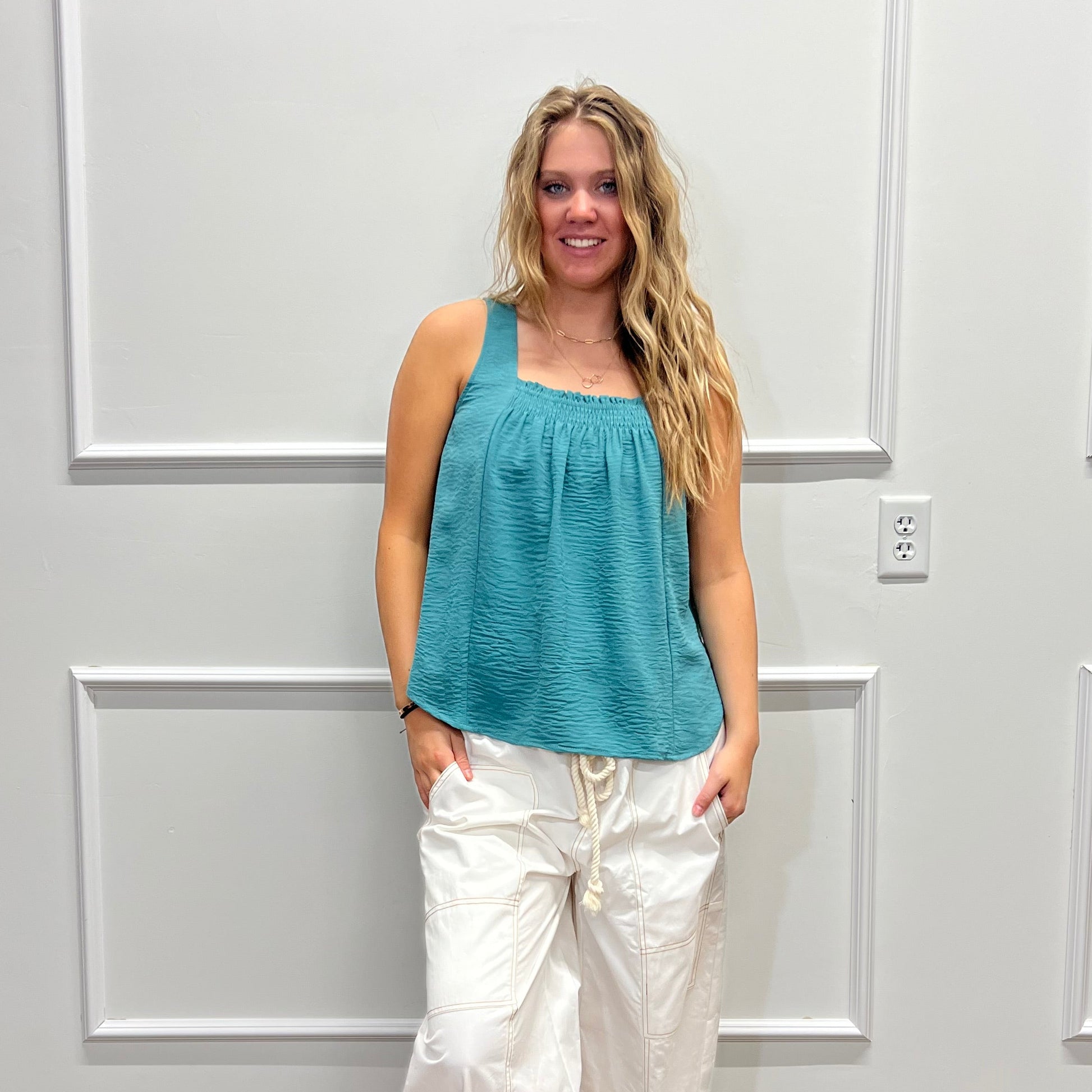 A woman stands indoors against a white paneled wall, wearing a sleeveless teal top and loose white pants with dark sandals. The photo is rotated sideways.