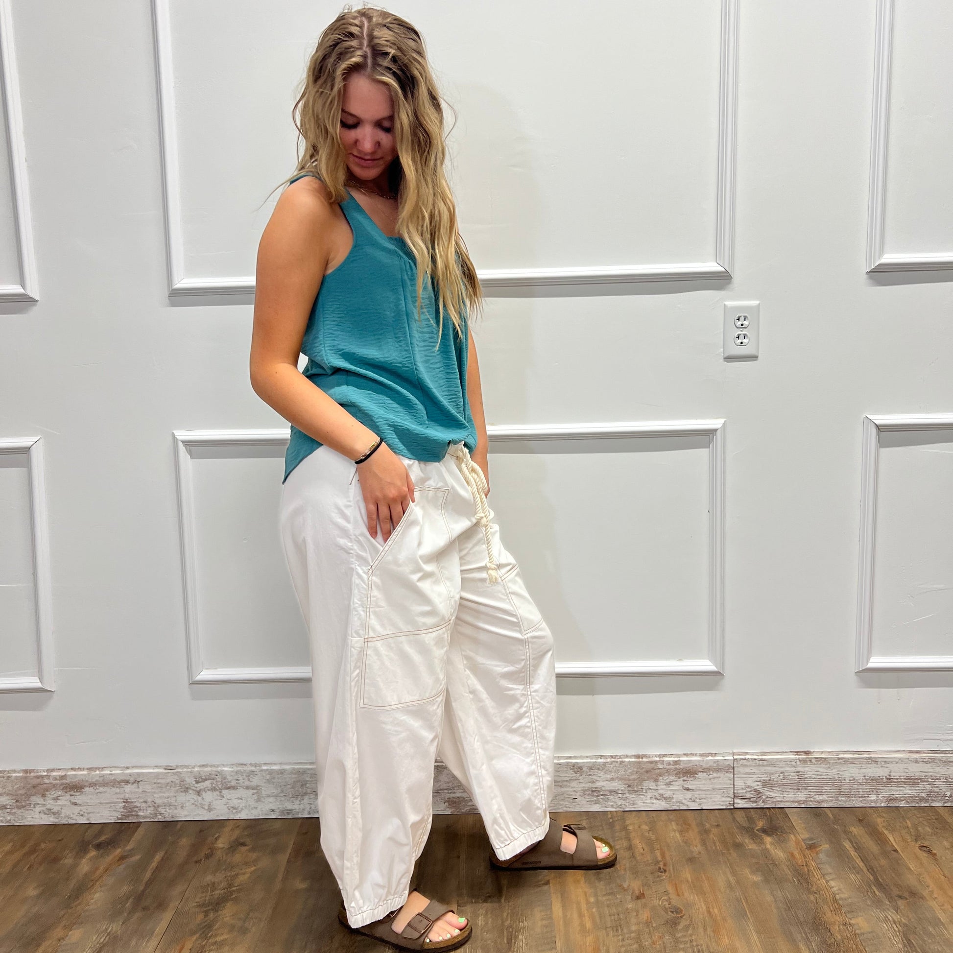 A woman wearing a teal sleeveless top and white wide-leg pants, standing in front of a white paneled wall with a wood floor visible on the right.