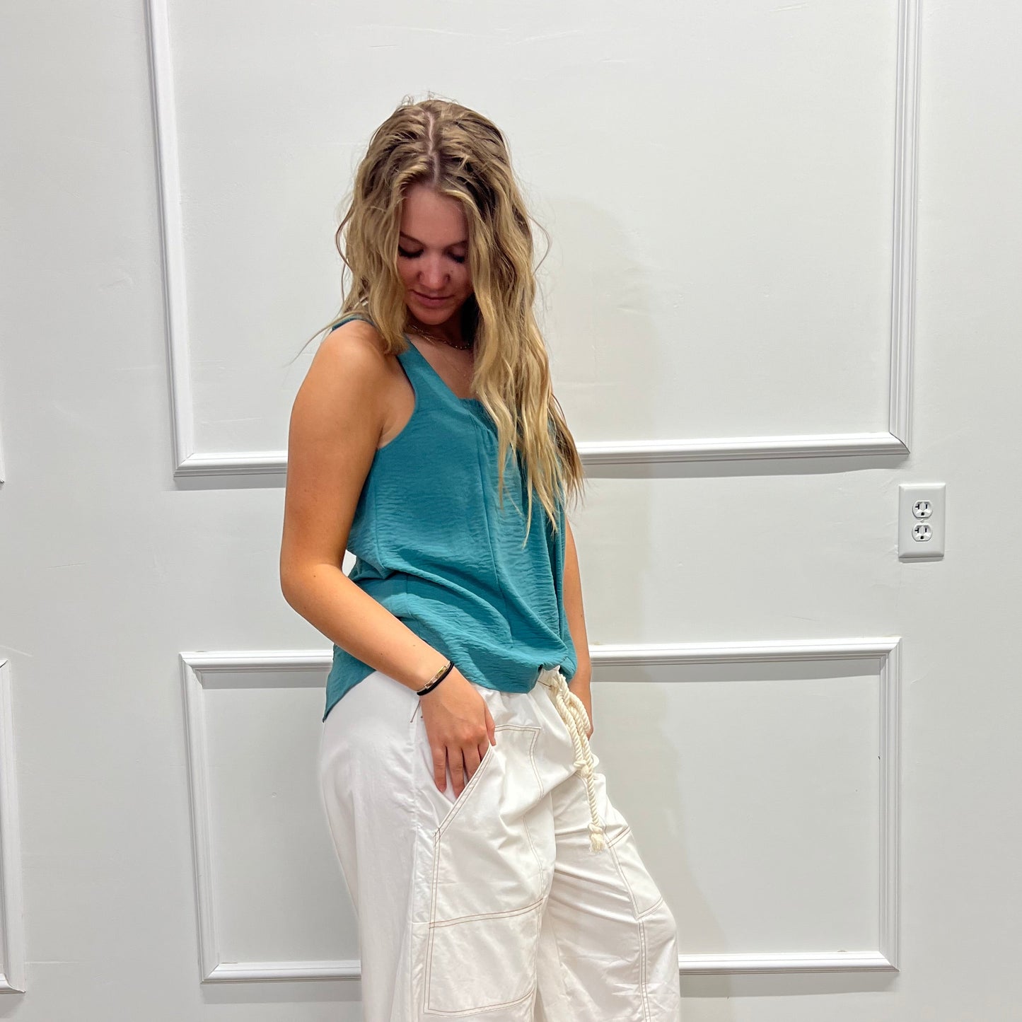 A woman wearing a teal sleeveless top and white wide-leg pants, standing in front of a white paneled wall with a wood floor visible on the right.