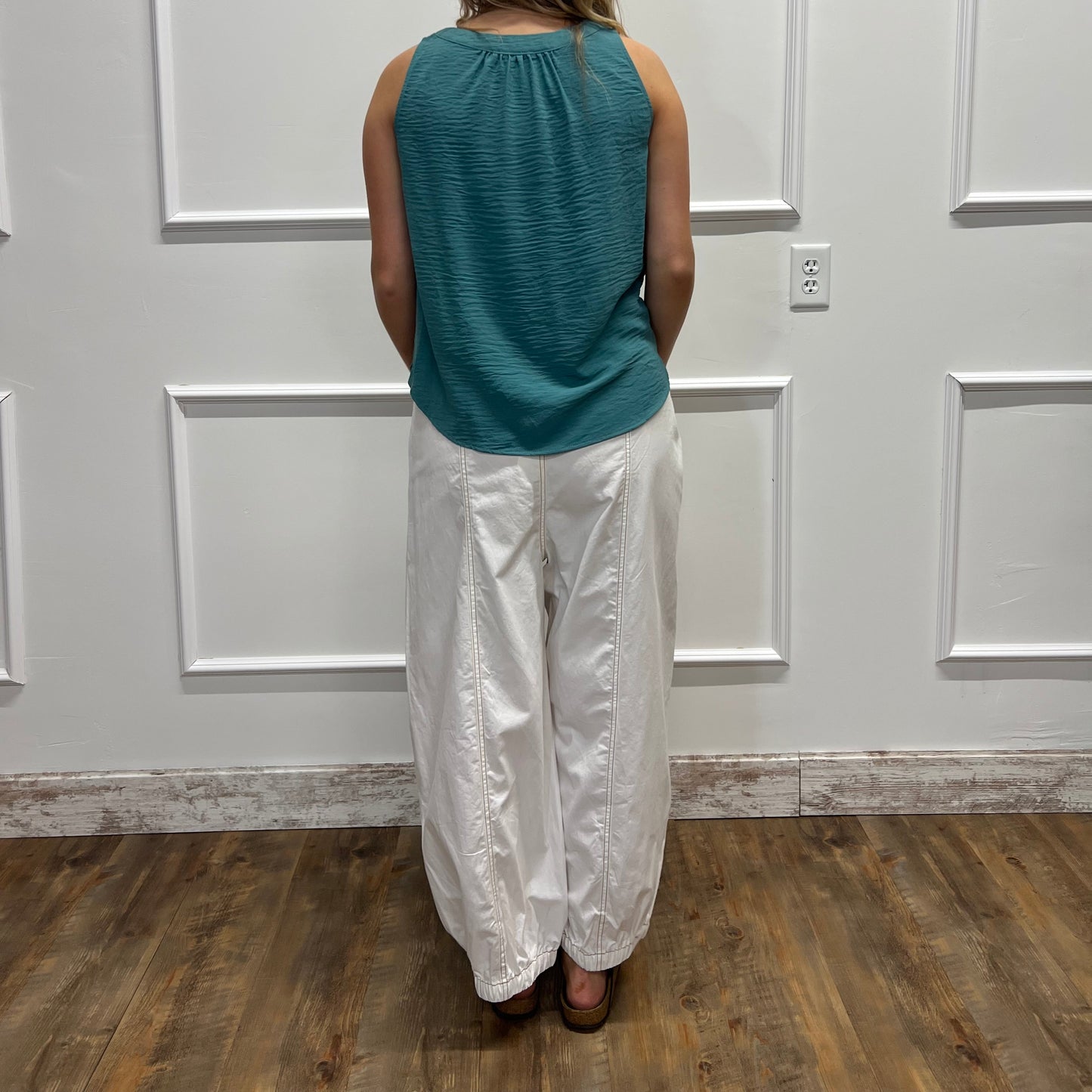 A woman stands with her back to the camera in front of a white paneled door, wearing a teal ribbed sleeveless top and shiny white pants, with a wood floor visible on the right.