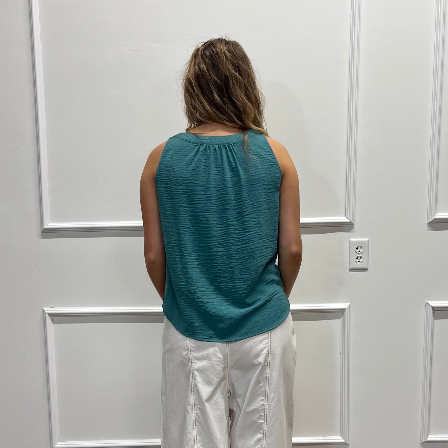 A woman stands with her back to the camera in front of a white paneled door, wearing a teal ribbed sleeveless top and shiny white pants, with a wood floor visible on the right.