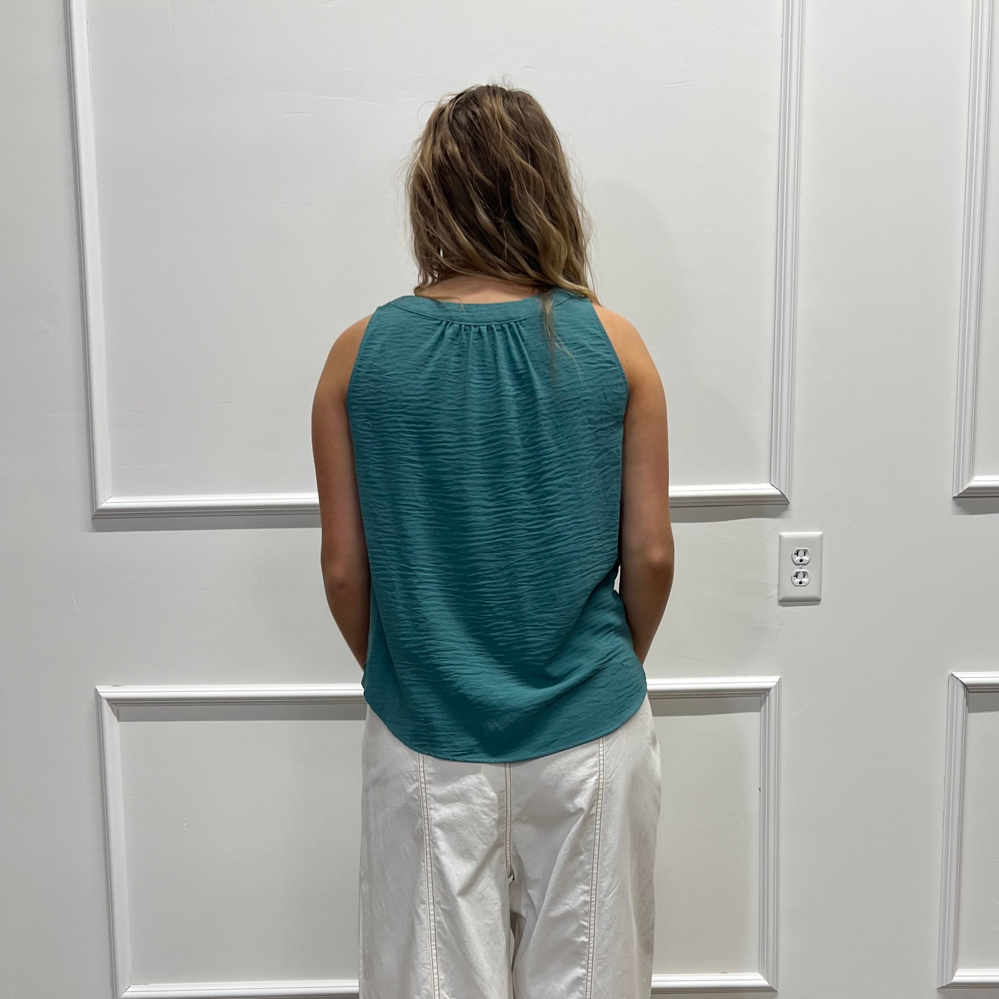 A woman stands with her back to the camera in front of a white paneled door, wearing a teal ribbed sleeveless top and shiny white pants, with a wood floor visible on the right.