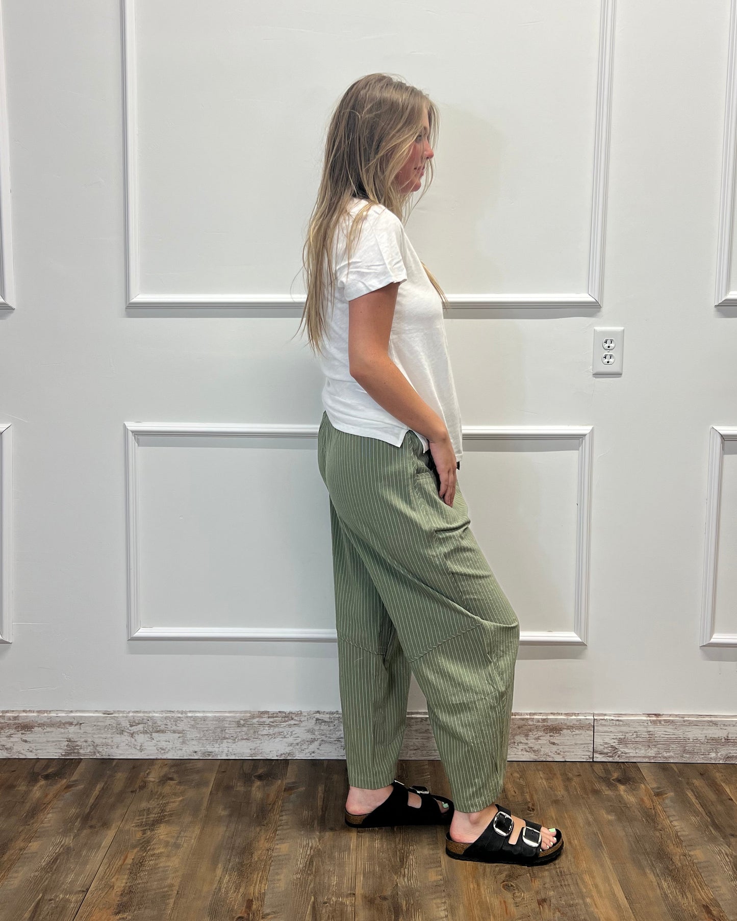 A woman wearing sage green wide-leg trousers, a white short-sleeve top, and black slide sandals stands in profile against a white paneled wall on a wood floor.