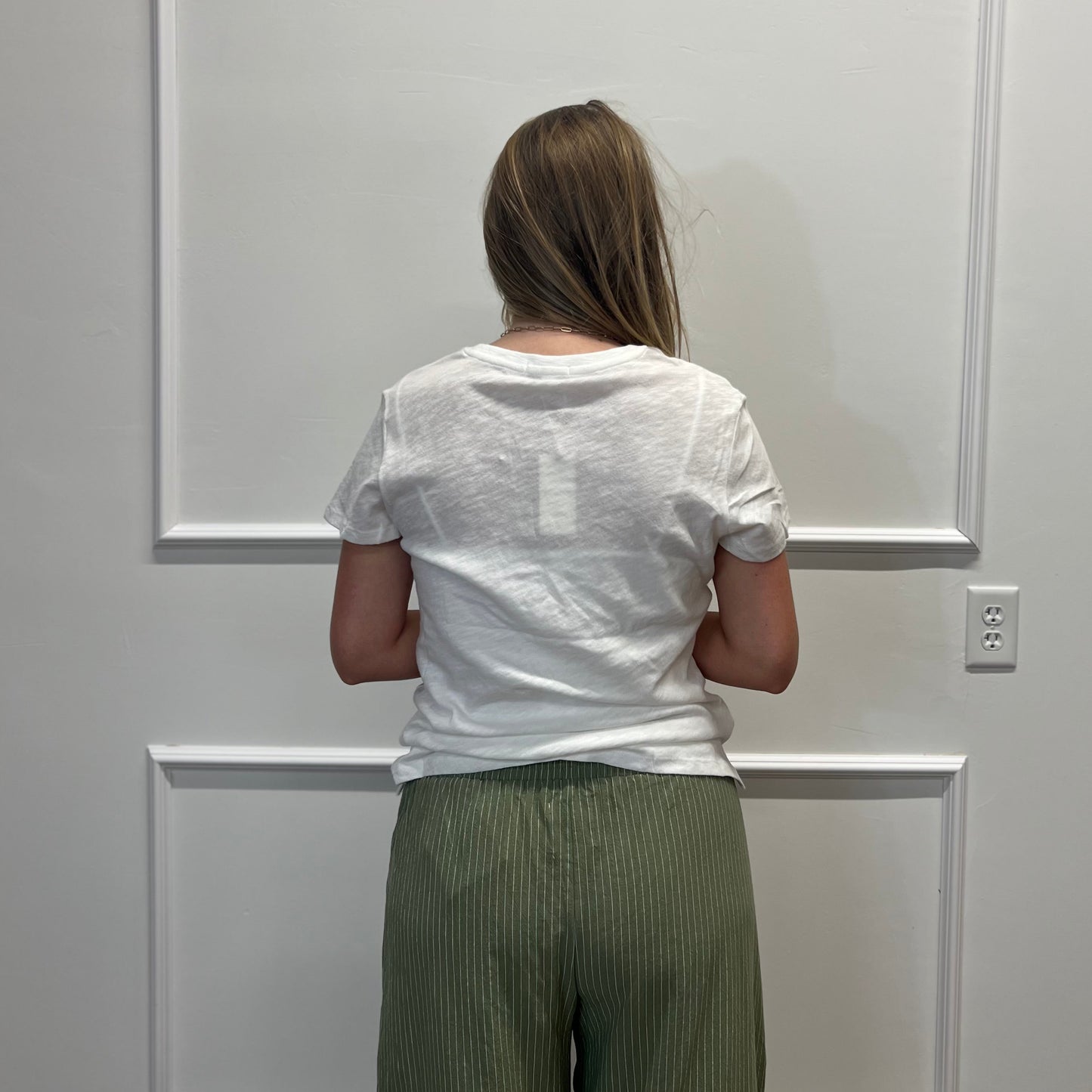 A woman viewed from behind wearing a white short-sleeve t-shirt and olive green pleated wide-leg pants, standing in front of a white paneled wall.