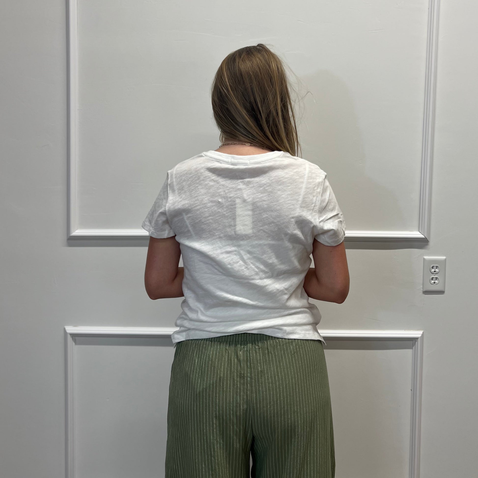 A woman viewed from behind wearing a white short-sleeve t-shirt and olive green pleated wide-leg pants, standing in front of a white paneled wall.