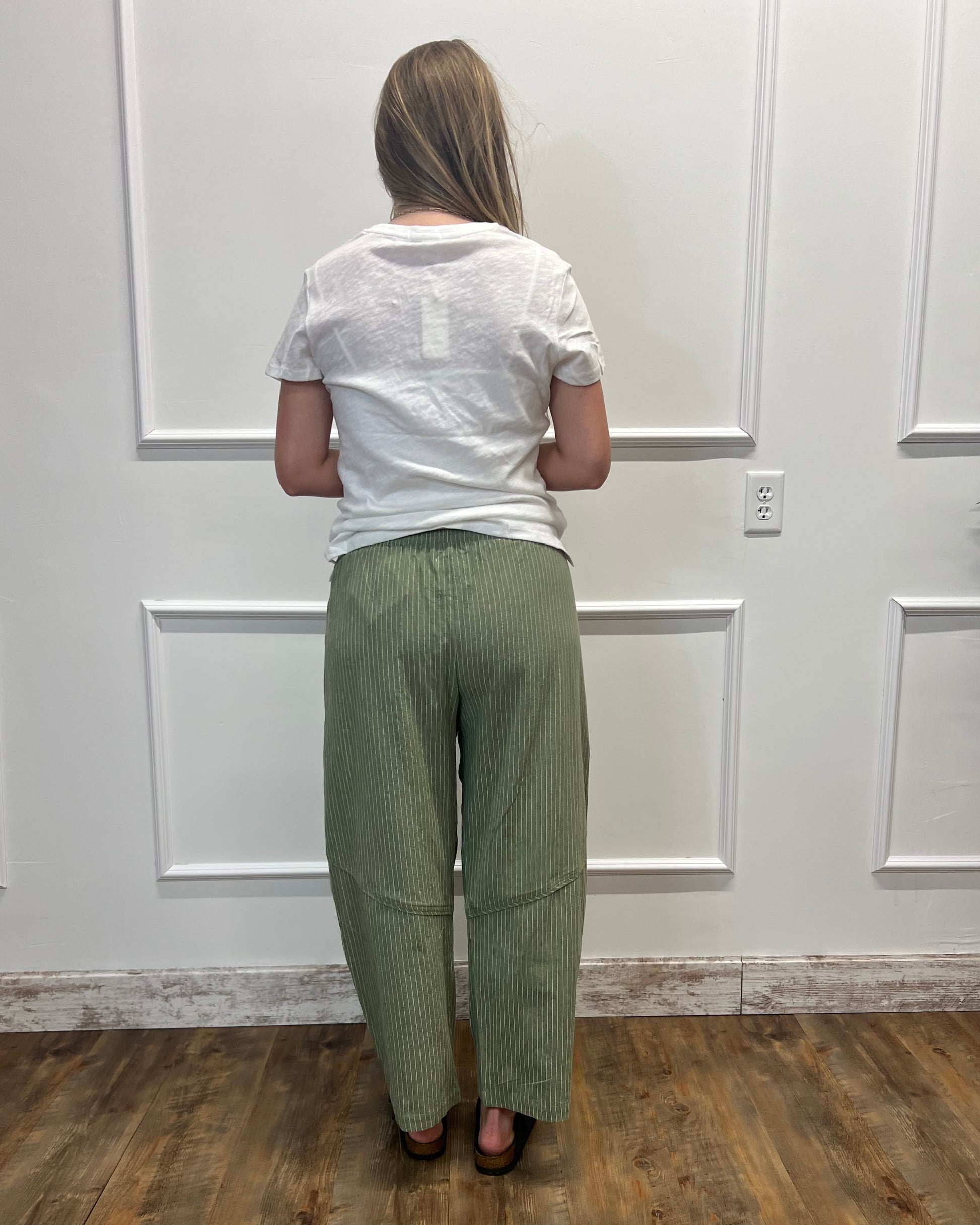 A woman viewed from behind wearing a white t-shirt and sage green wide-leg corduroy pants, standing on a wood floor against a white paneled wall.