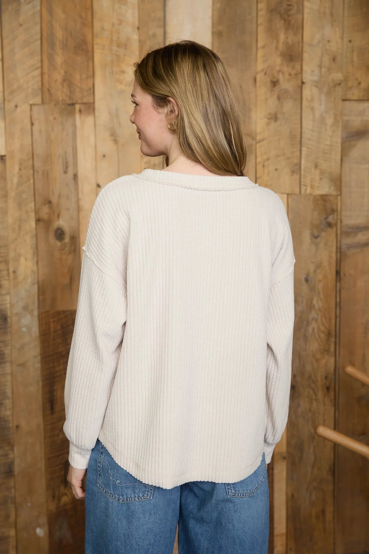 Woman wearing a beige sweater and blue jeans against a wooden wall.