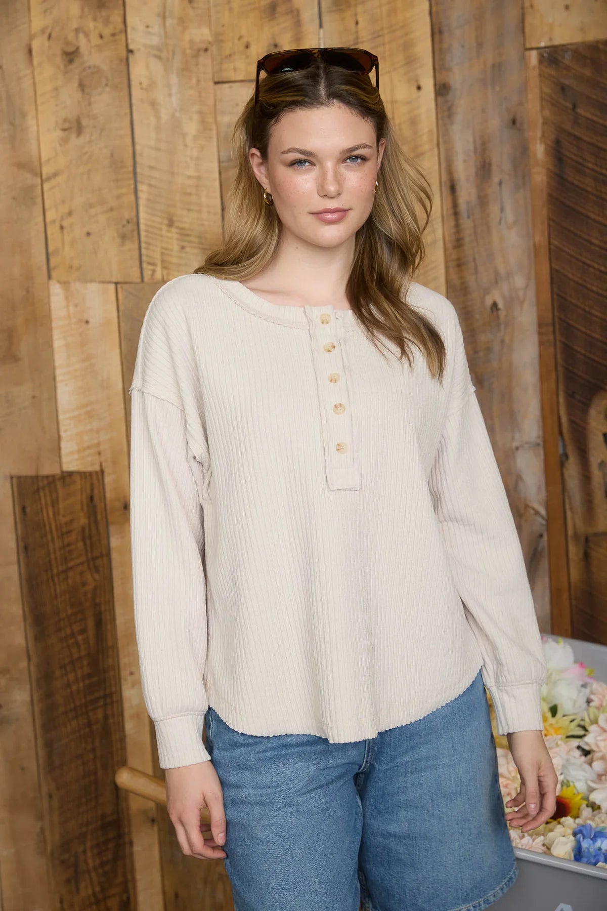 Woman wearing a beige blouse with buttons and blue jeans against a wooden wall.