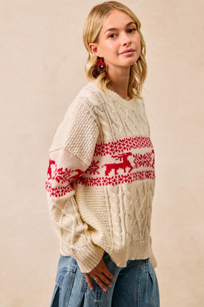 A woman wearing a cream cable-knit sweater with a red Nordic-style pattern across the chest and sleeves, paired with blue jeans, set against a plain beige background.