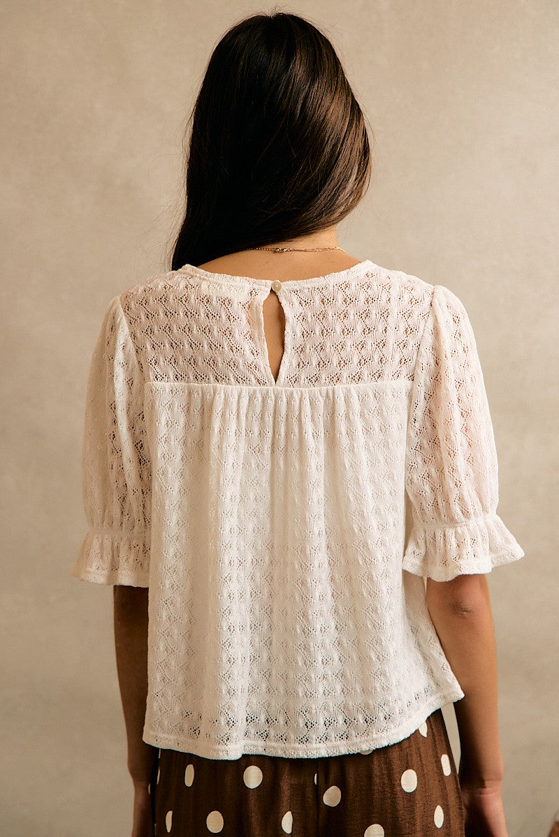 A woman viewed from behind wearing a cream-colored, short-sleeved lace blouse with a keyhole back cutout and brown polka-dot skirt against a neutral beige background.